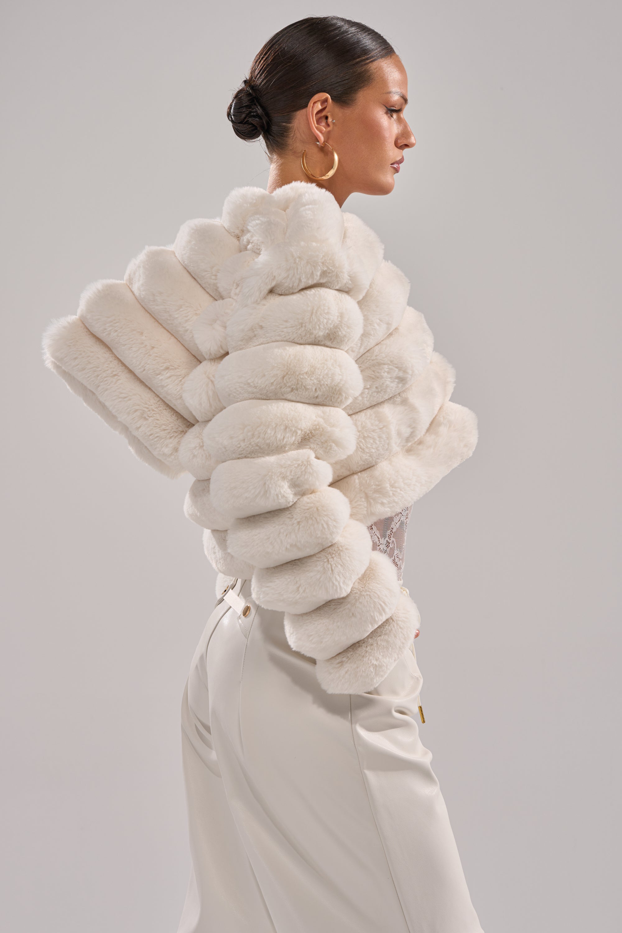 A woman with slicked-back hair models the POPCORN FAUX FUR IN WHITE jacket and high-waisted cream pants, standing in profile against a light gray background—a chic winter fashion statement. 5