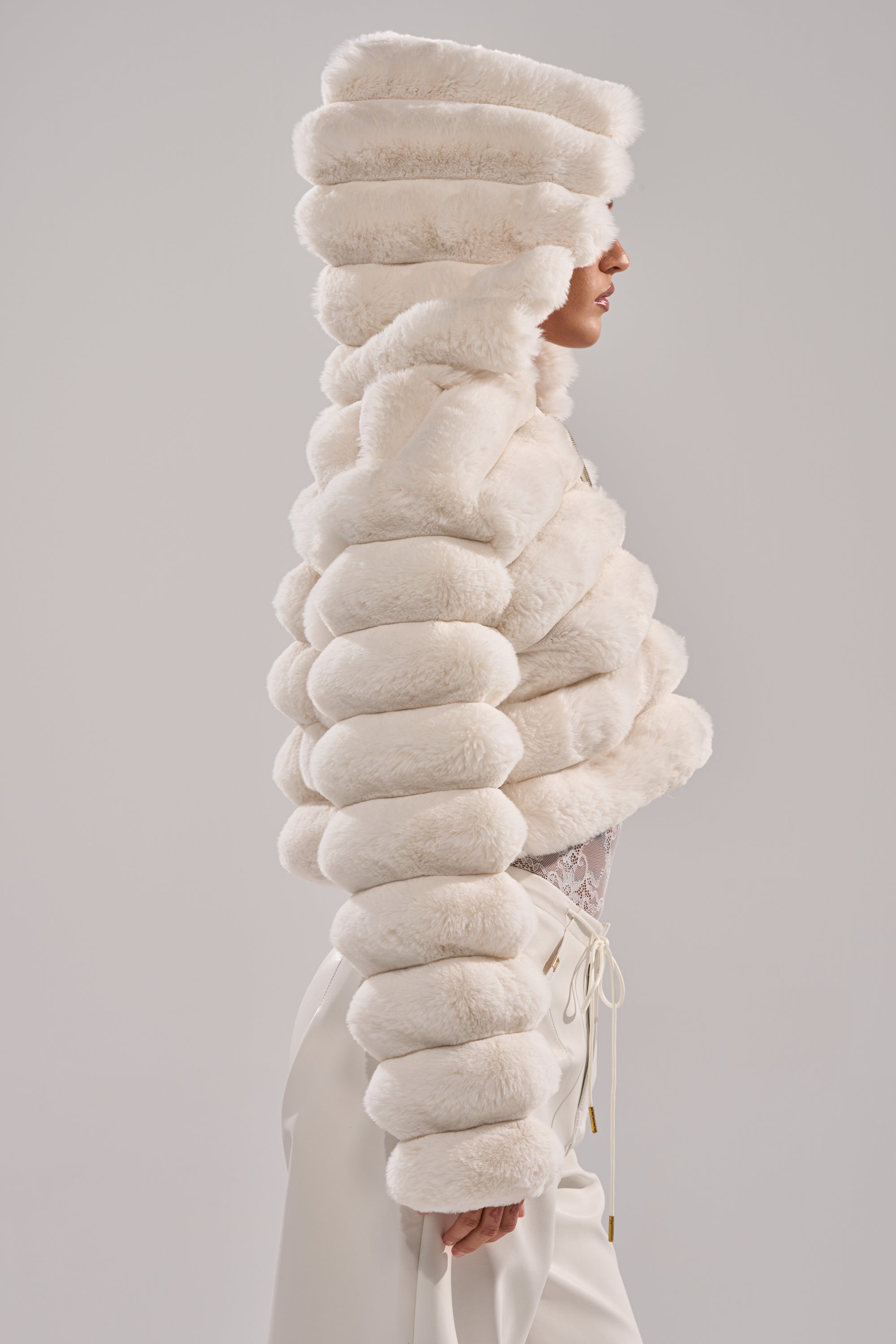 Wearing the POPCORN FAUX FUR IN WHITE, a person in profile showcases its dramatic, segmented design from hood to sleeves, paired with white pants against a neutral background for a chic winter look. 6