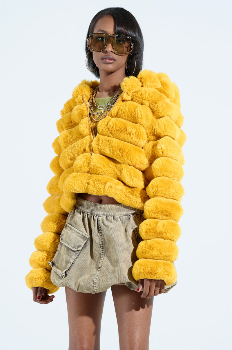 Wearing the POPCORN FAUX FUR IN YELLOW and a beige skirt, accessorized with oversized sunglasses and layered necklaces against a plain white background.