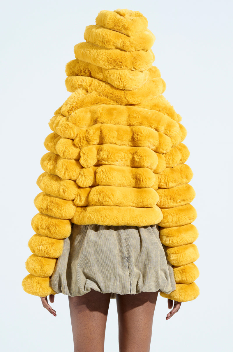 A person faces away from the camera in a bright yellow POPCORN FAUX FUR jacket with oversized spiral-like padding and loose tan shorts, set against a plain light blue background.