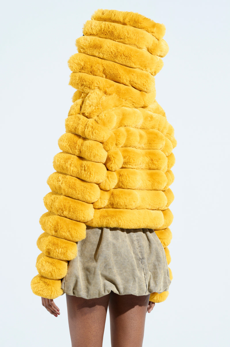 A person is turned away, wearing the POPCORN FAUX FUR IN YELLOW, a bright yellow puffy cropped jacket with a hood. They pair it with tan, textured shorts against a plain light blue background.