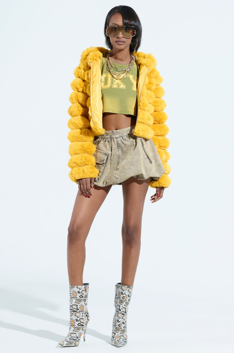 A person confidently wears the POPCORN FAUX FUR IN YELLOW jacket over a green "01" crop top, paired with high-waisted shorts, sunglasses, necklaces, and patterned high-heeled boots.