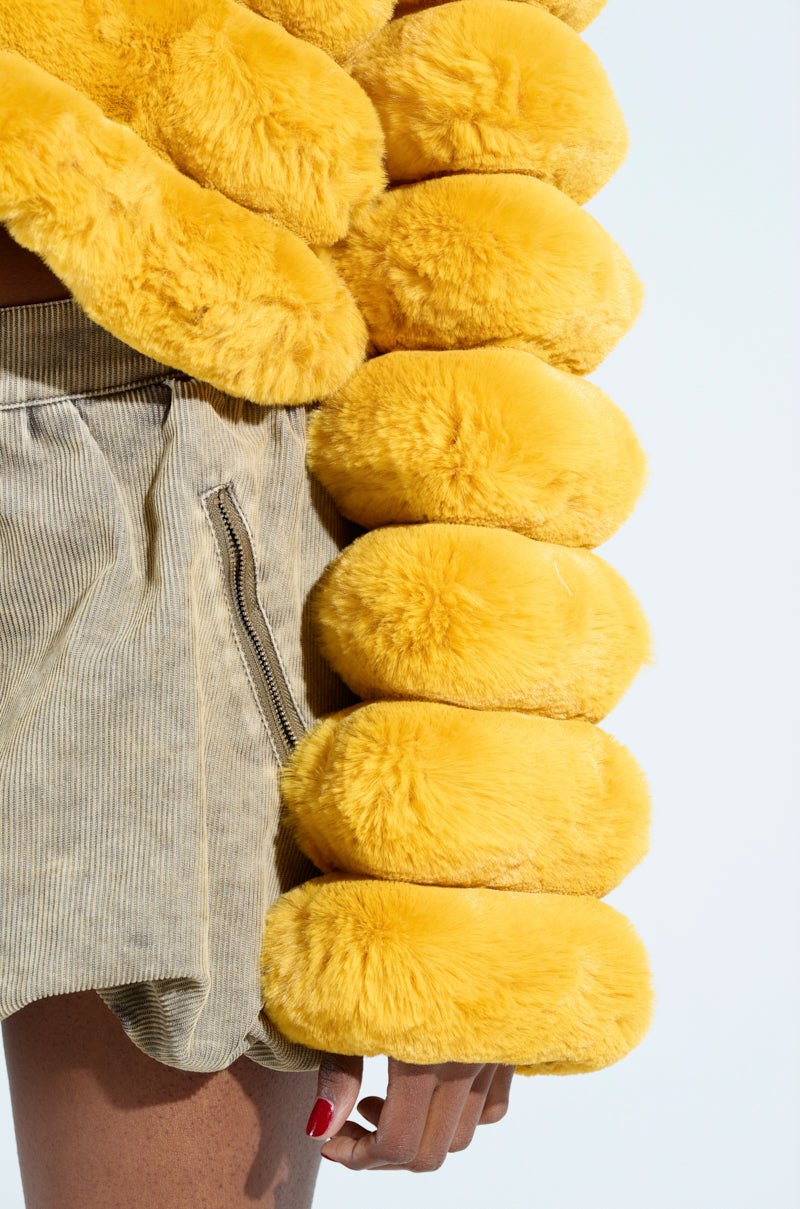 Wearing the POPCORN FAUX FUR IN YELLOW, featuring large rounded shapes along the sleeves, paired with beige corduroy shorts, this ensemble stands out against a light background with a hint of the person's hand visible.