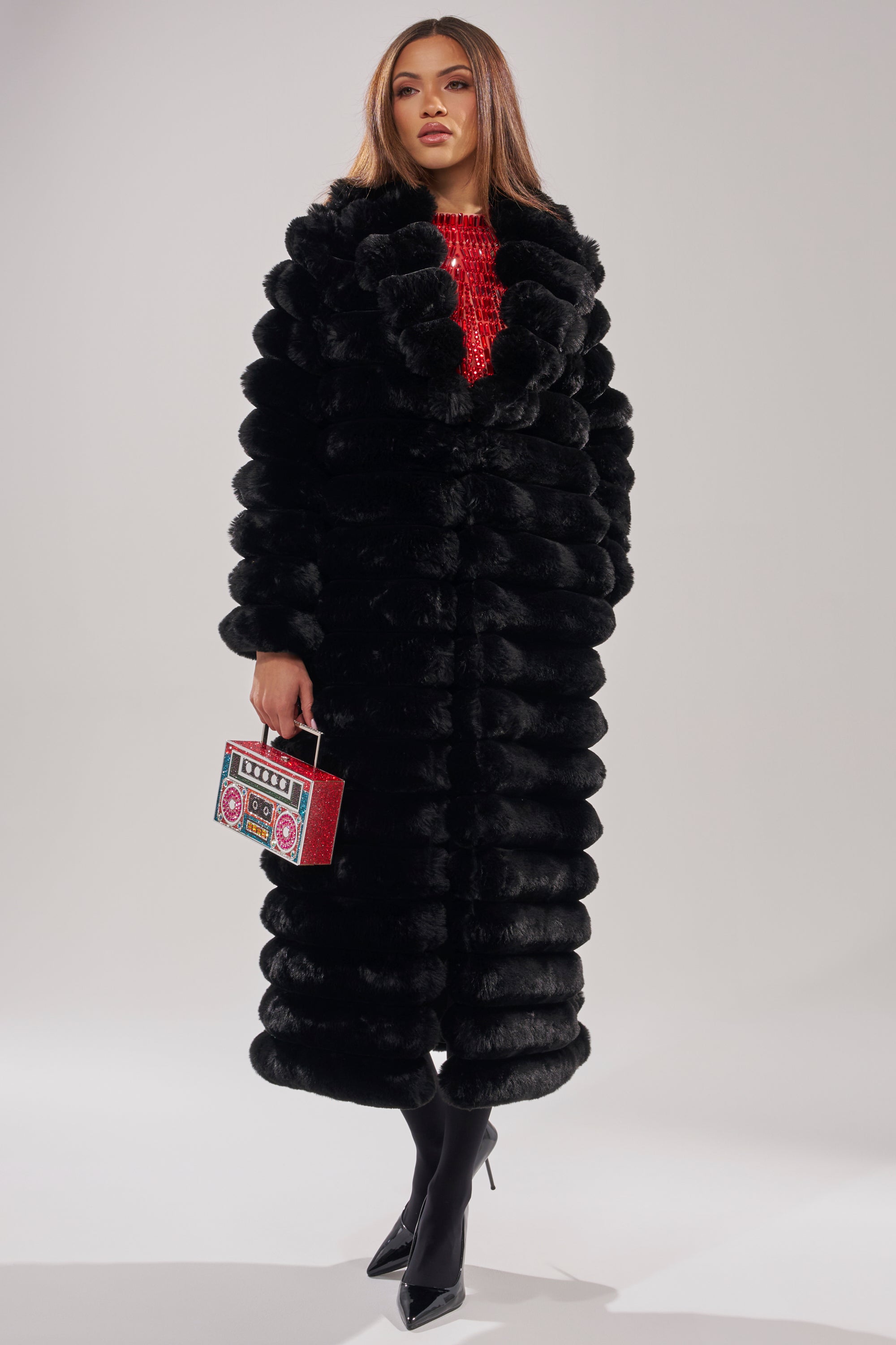 A woman models the MAXI POPCORN FAUX FUR IN BLACK over a red top, holding a vibrant box-shaped handbag. She has straight brown hair, wears black heels, and is posed against a plain light background.