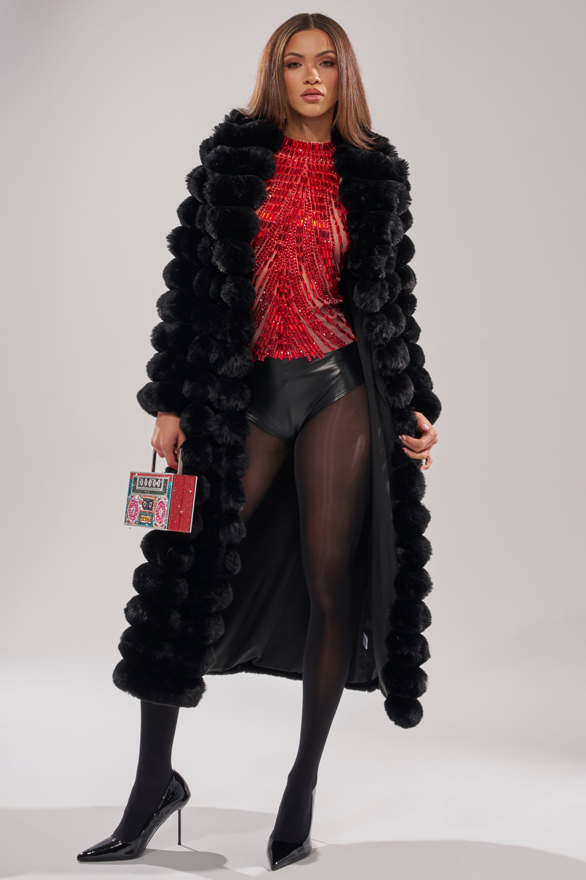 A woman stands confidently in the MAXI POPCORN FAUX FUR IN BLACK coat, styled with a red beaded top, black shorts, sheer tights, high heels, and a colorful clutch against a plain background.