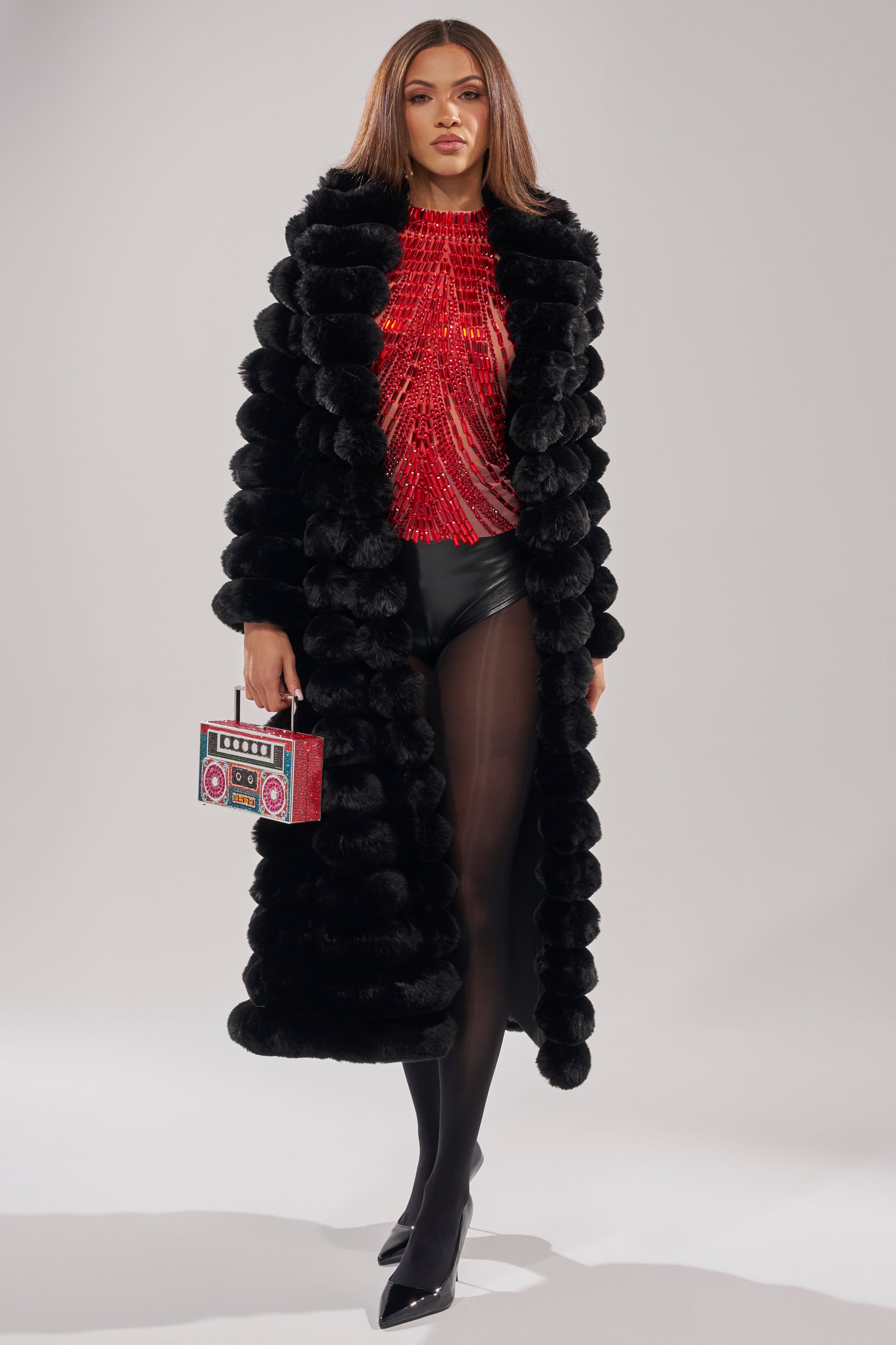 A woman exudes confidence in the MAXI POPCORN FAUX FUR IN BLACK, styled over a red textured top and black shorts. She wears black tights, pointed-toe heels, and carries a small handbag shaped like a vintage boombox.