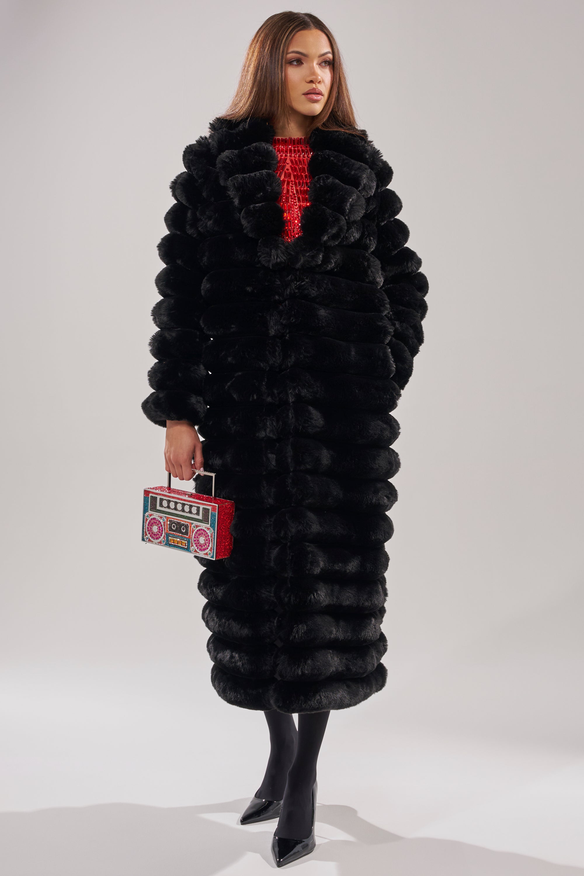 A woman wears the MAXI POPCORN FAUX FUR IN BLACK coat over a red top, black tights, and pointed heels, holding a red boombox-shaped purse against a plain light gray background.