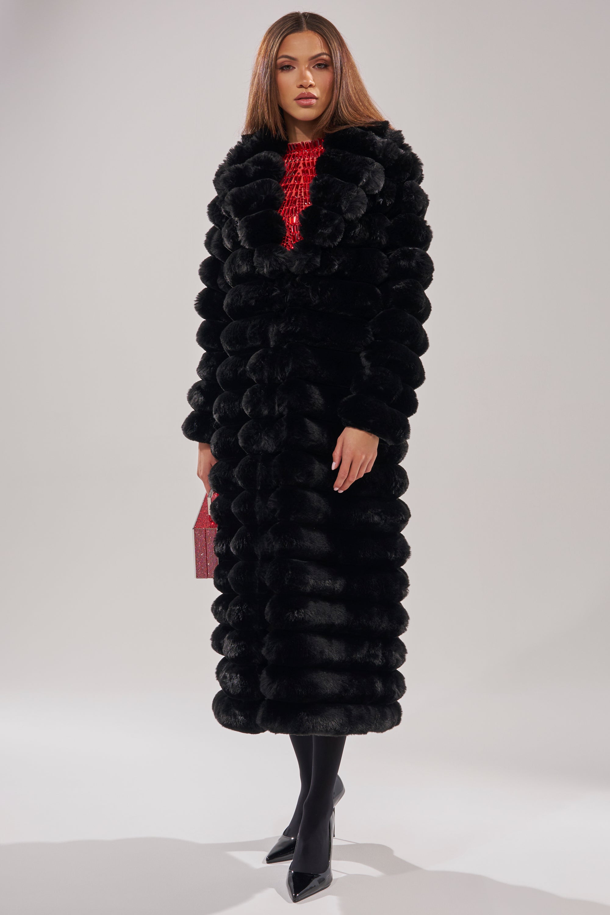 A woman wears the MAXI POPCORN FAUX FUR IN BLACK coat over a red top, black tights, and pointed-toe heels, holding a small red clutch against a plain light background.