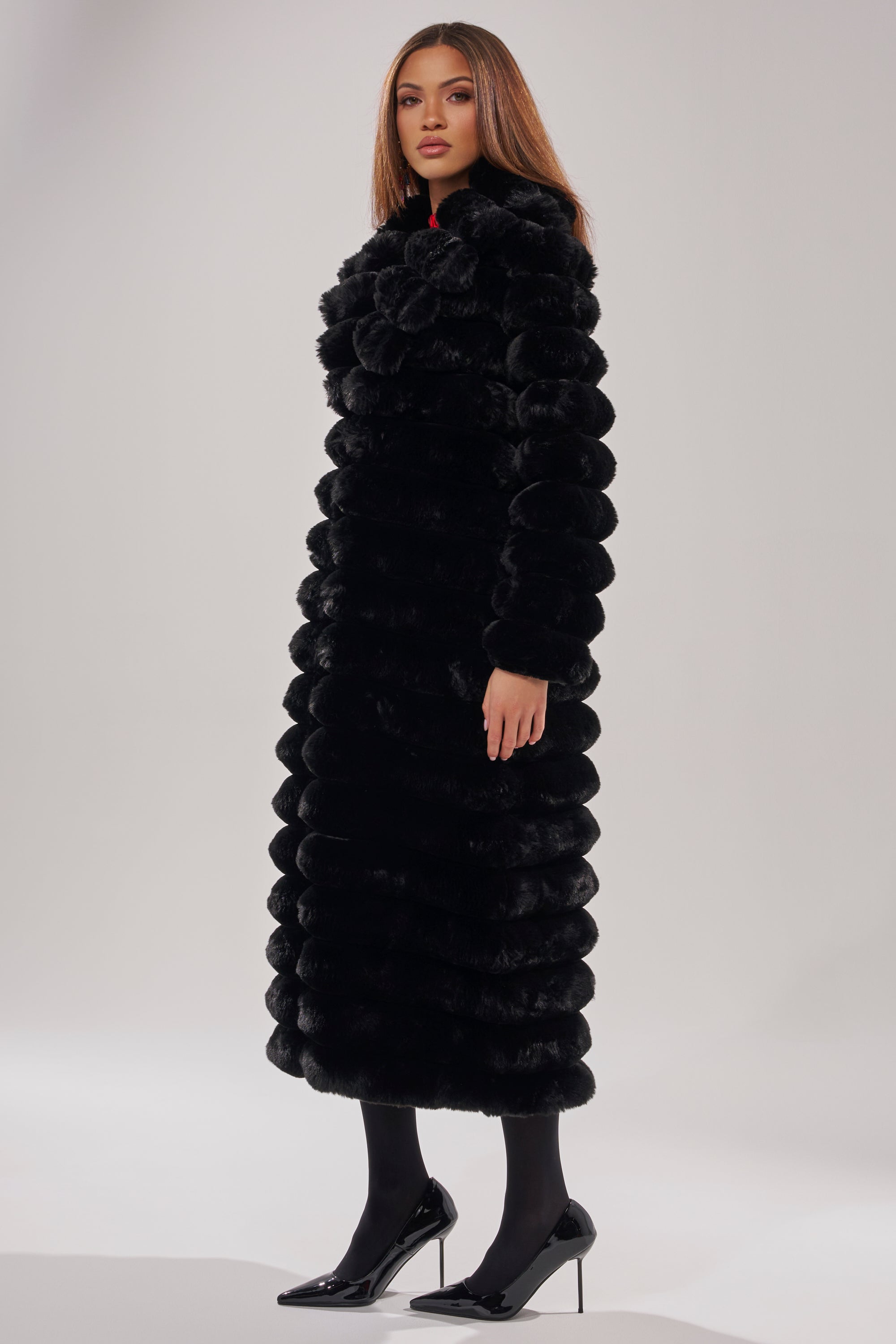 A woman with straight brown hair and a neutral expression stands in a studio wearing the MAXI POPCORN FAUX FUR IN BLACK coat, paired with black tights and high-heeled shoes.