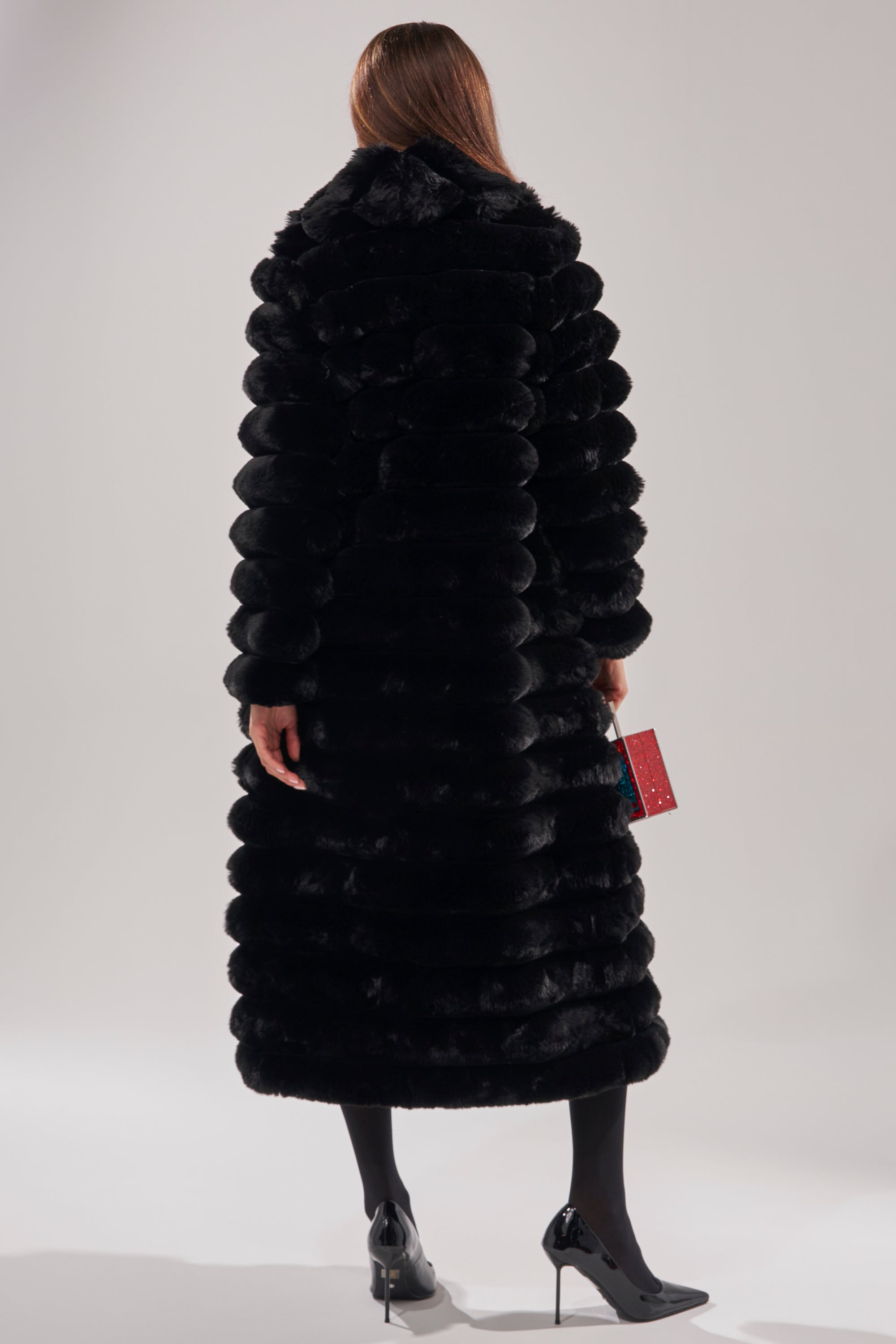 A person with long brown hair is seen from behind, wearing the MAXI POPCORN FAUX FUR IN BLACK and black high-heeled shoes, holding a small red clutch purse against a plain light background.