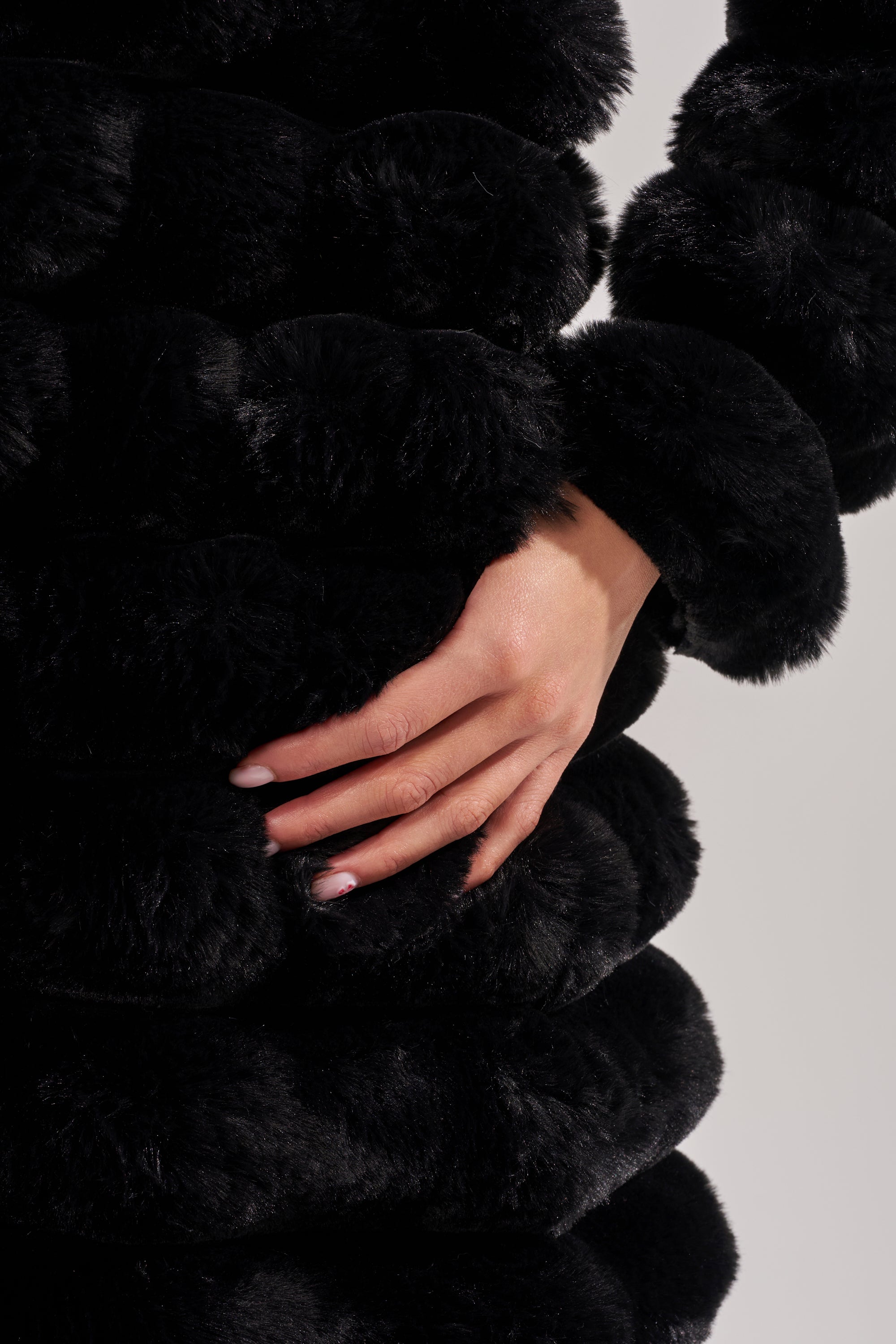 A close-up of a hand resting on the waist, wearing the MAXI POPCORN FAUX FUR IN BLACK with horizontal, fluffy ridges. Fingernails are neatly manicured in a natural shade.