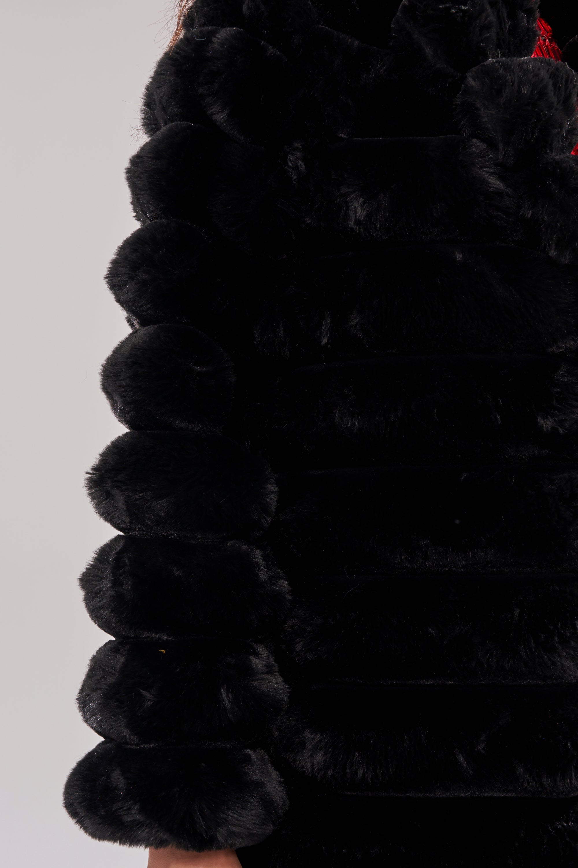 Close-up of a person wearing the MAXI POPCORN FAUX FUR IN BLACK, featuring round, plush sections and a visible hand. The textured black coat stands out against a plain light gray background.