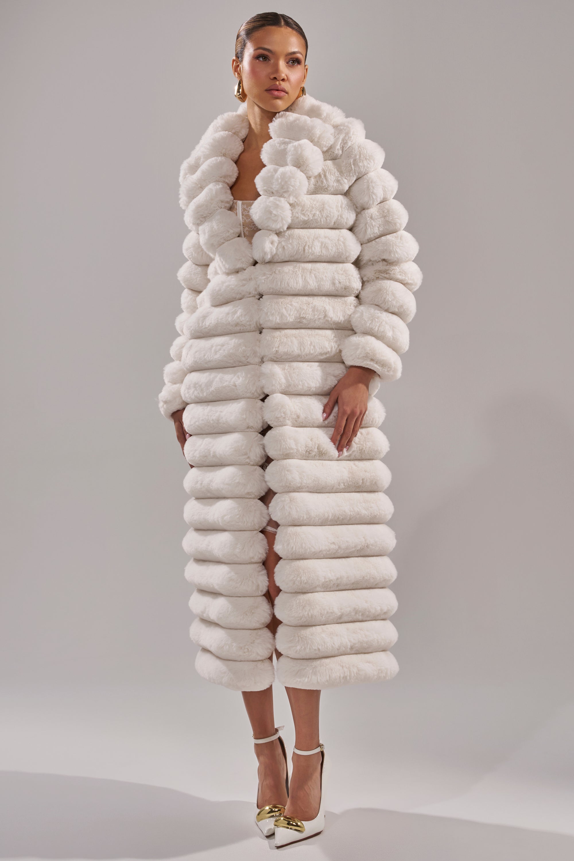 A woman models the MAXI POPCORN FAUX FUR IN WHITE coat with thick horizontal segments, paired with white heels featuring gold accents and hoop earrings, set against a plain light gray background.