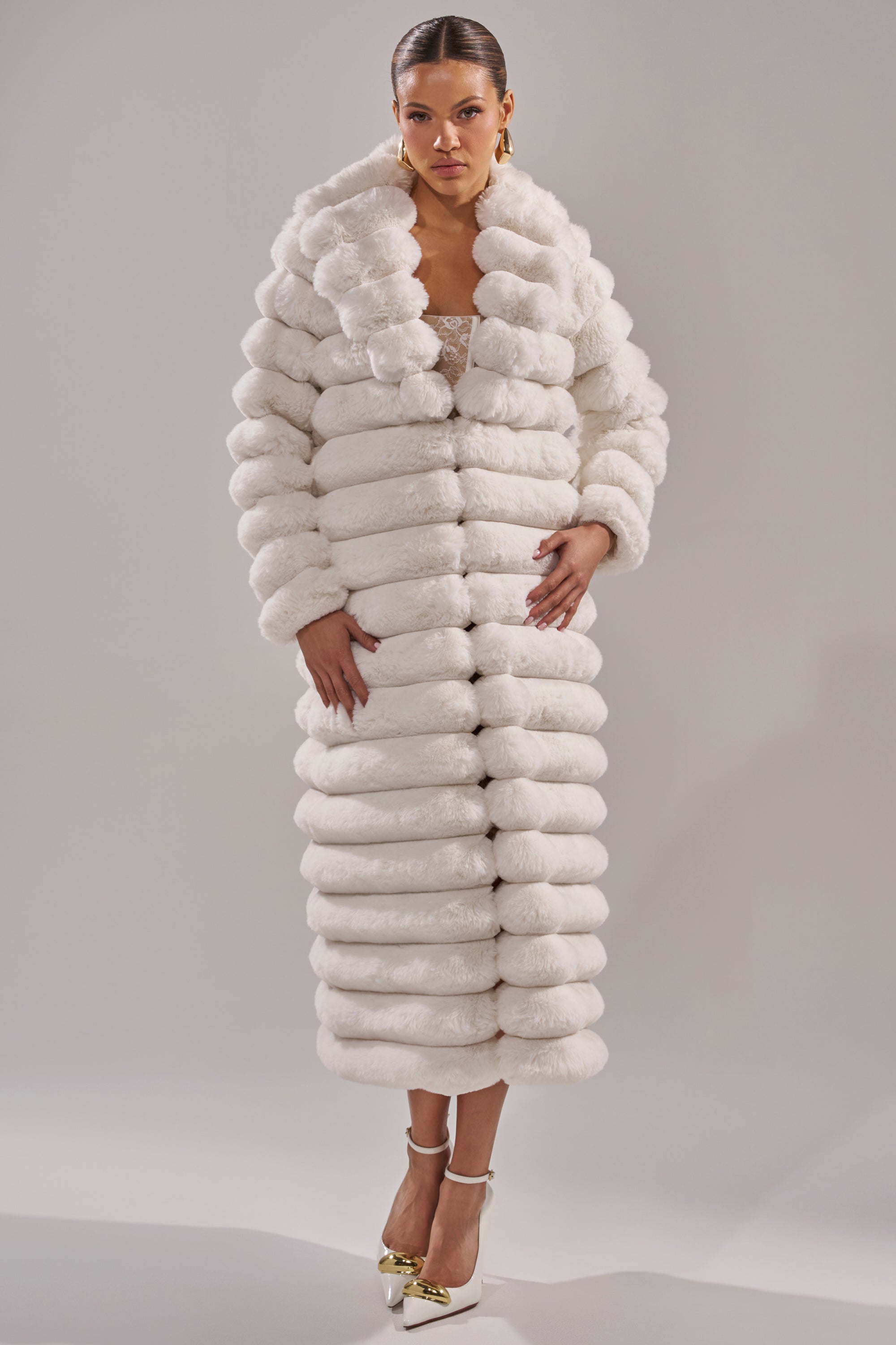 A woman with slicked-back hair, gold earrings, and white high heels with gold details wears the MAXI POPCORN FAUX FUR IN WHITE coat, featuring luxurious thick horizontal bands, standing against a plain background.
