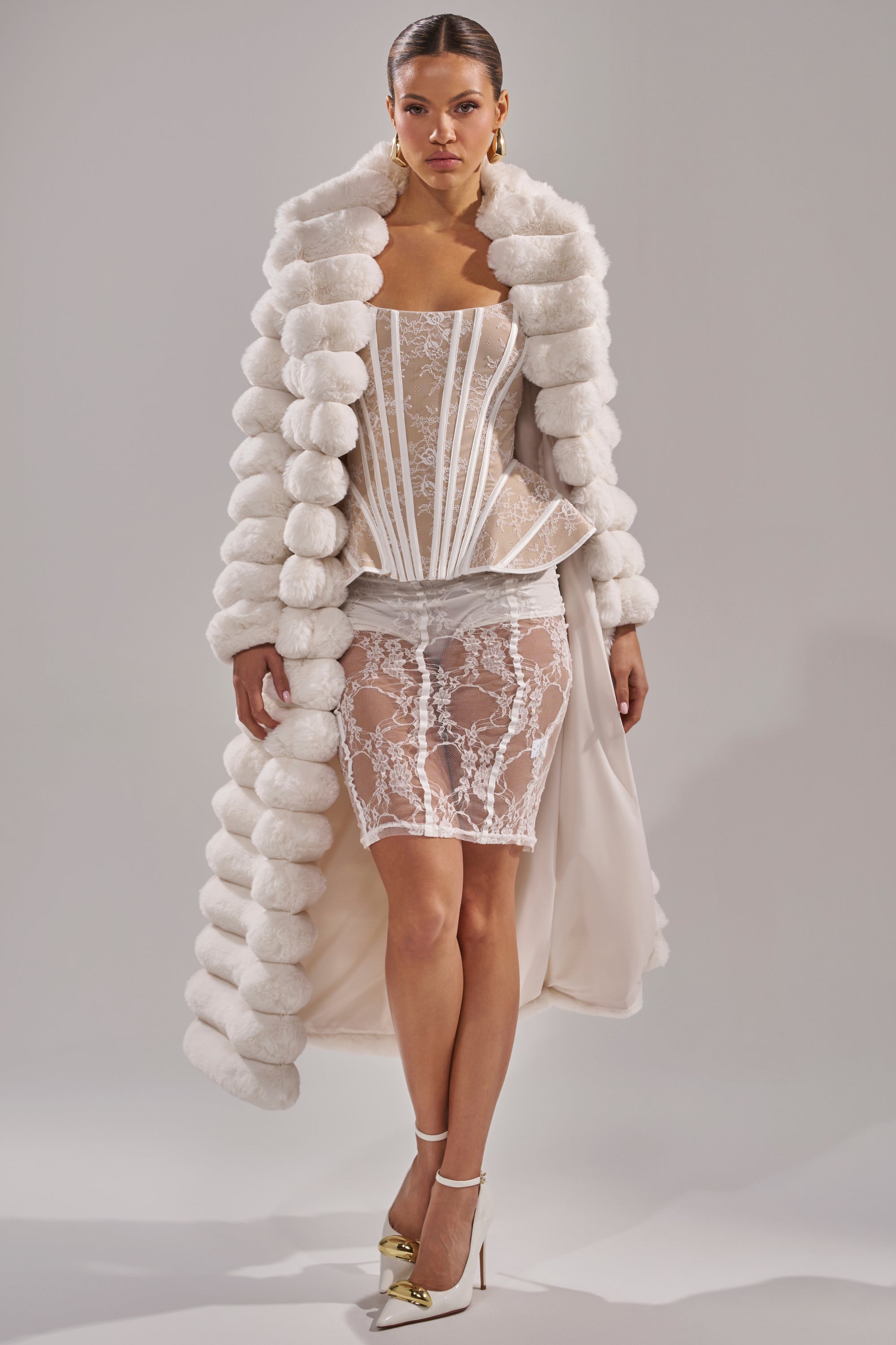 A woman models a white lace corset top, sheer skirt, and the MAXI POPCORN FAUX FUR IN WHITE coat with white heels featuring gold accents, set against a simple gray backdrop.