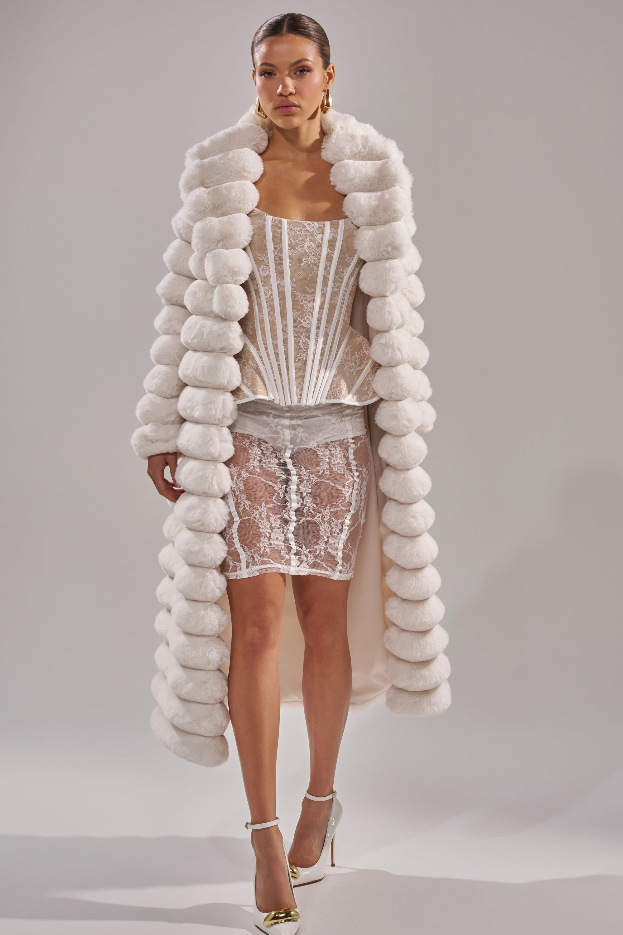 A woman models a sheer white lace corset dress with a structured bodice and short hem, styled with the MAXI POPCORN FAUX FUR IN WHITE—a dramatic puffy ribbed maxi coat. She completes the look with white heels and gold earrings, posing confidently.
