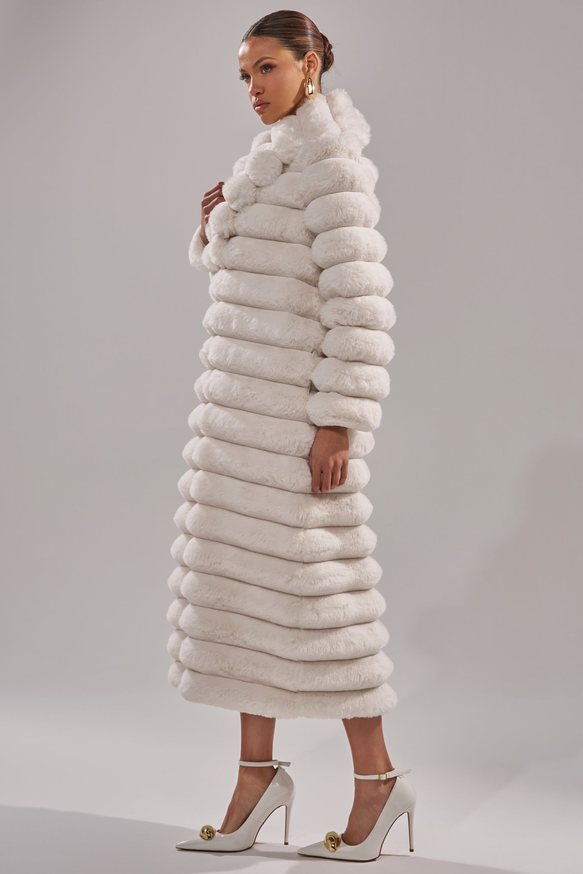 A woman poses in the MAXI POPCORN FAUX FUR IN WHITE, a luxurious ribbed white maxi coat, styled with white heels and statement gold earrings against a minimal light background.