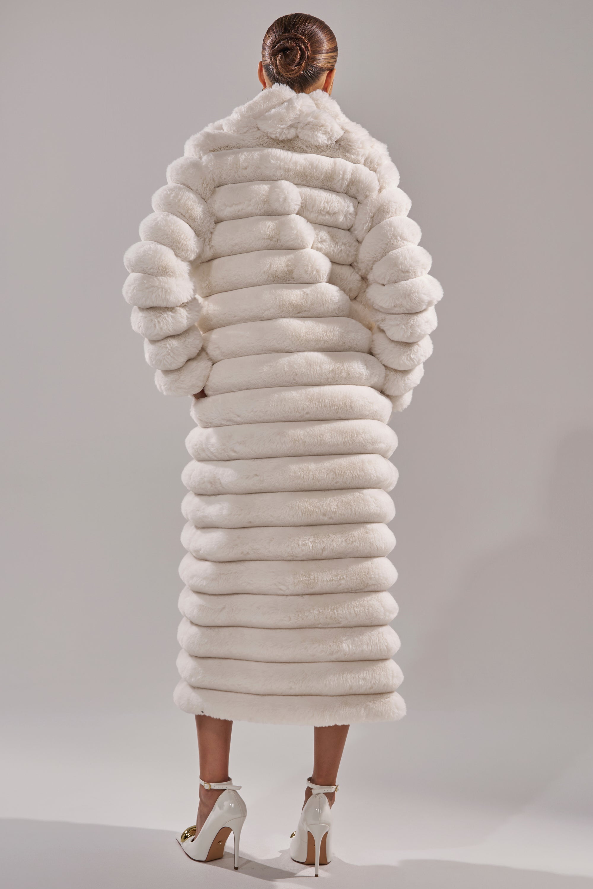 A person with a neat bun faces away, wearing the MAXI POPCORN FAUX FUR IN WHITE and white high heels, set against a plain neutral background.