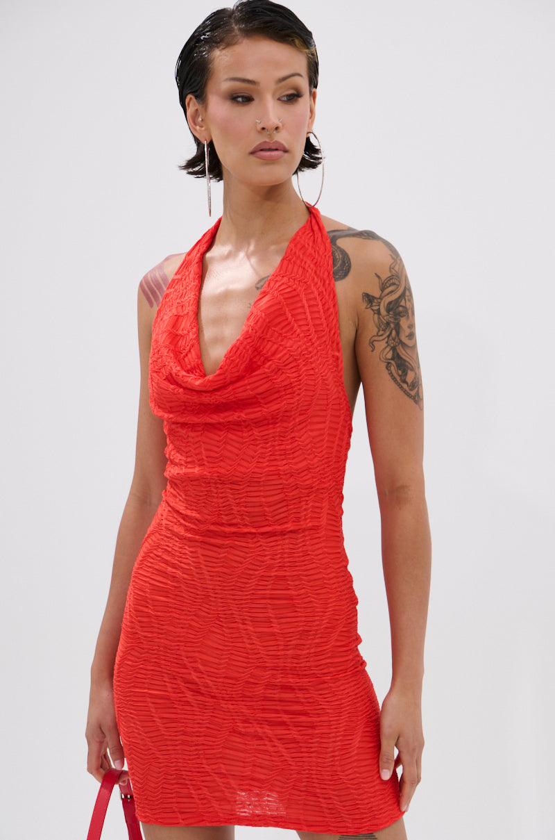 A woman with short dark hair wears the ALWAYS BEST DRESSED MINI DRESS IN RED, a sleeveless, textured mini dress with a draped neckline and fitted silhouette. She has an arm tattoo, holds a pink handbag, and looks to the side against a plain background.