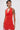 A woman with short dark hair wears the ALWAYS BEST DRESSED MINI DRESS IN RED, a sleeveless, textured mini dress with a draped neckline and fitted silhouette. She has an arm tattoo, holds a pink handbag, and looks to the side against a plain background.