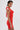 A woman with short dark hair and a tattoo on her upper arm wears the ALWAYS BEST DRESSED MINI DRESS IN RED—a curve-hugging, sleeveless halter-neck mini dress—posing sideways against a plain white background with large hoop earrings.