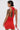 Wearing the ALWAYS BEST DRESSED MINI DRESS IN RED—a red, open-back mini that hugs curves—a person with short dark hair, large hoop earrings, and visible tattoos turns away from the camera holding a matching red strap.