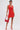 A person poses against a white background wearing the ALWAYS BEST DRESSED MINI DRESS IN RED, paired with red strappy heels and a matching handbag. Their hair is slicked back and several tattoos are visible.