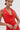 A person with short dark hair, hoop earrings, and tattoos models the ALWAYS BEST DRESSED MINI DRESS IN RED—a curve-hugging mini dress—standing with hands clasped and a neutral expression against a plain light background.