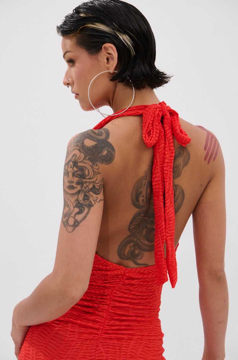 A person with short, slicked-back hair and hoop earrings models the ALWAYS BEST DRESSED MINI DRESS IN RED—a curve-hugging, textured mini dress with an open back—revealing tattoos on their upper arms and back against a plain background.