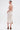 A woman with her hair in a bun wears the ALWAYS BEST DRESSED MIDI DRESS IN IVORY, a cream crochet open-back halter midi dress, paired with brown high-heeled sandals, standing against a plain white background.