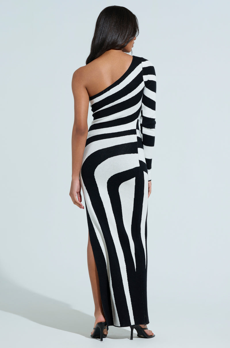 A woman with long hair stands with her back turned, highlighting a swirling striped pattern on her black and white STRIPED SINGLE SLEEVE SWEATER DRESS. Black heels add flair, while the side slit reveals its chic, maxi-length silhouette.