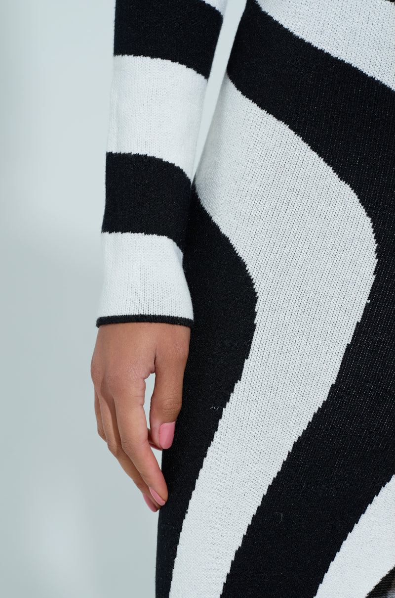 Someone sports a striking STRIPED SINGLE SLEEVE SWEATER DRESS with a swirling black and white pattern. The visible arm and part of the torso highlight its bold design, as the hand rests casually against a plain, light-colored background.