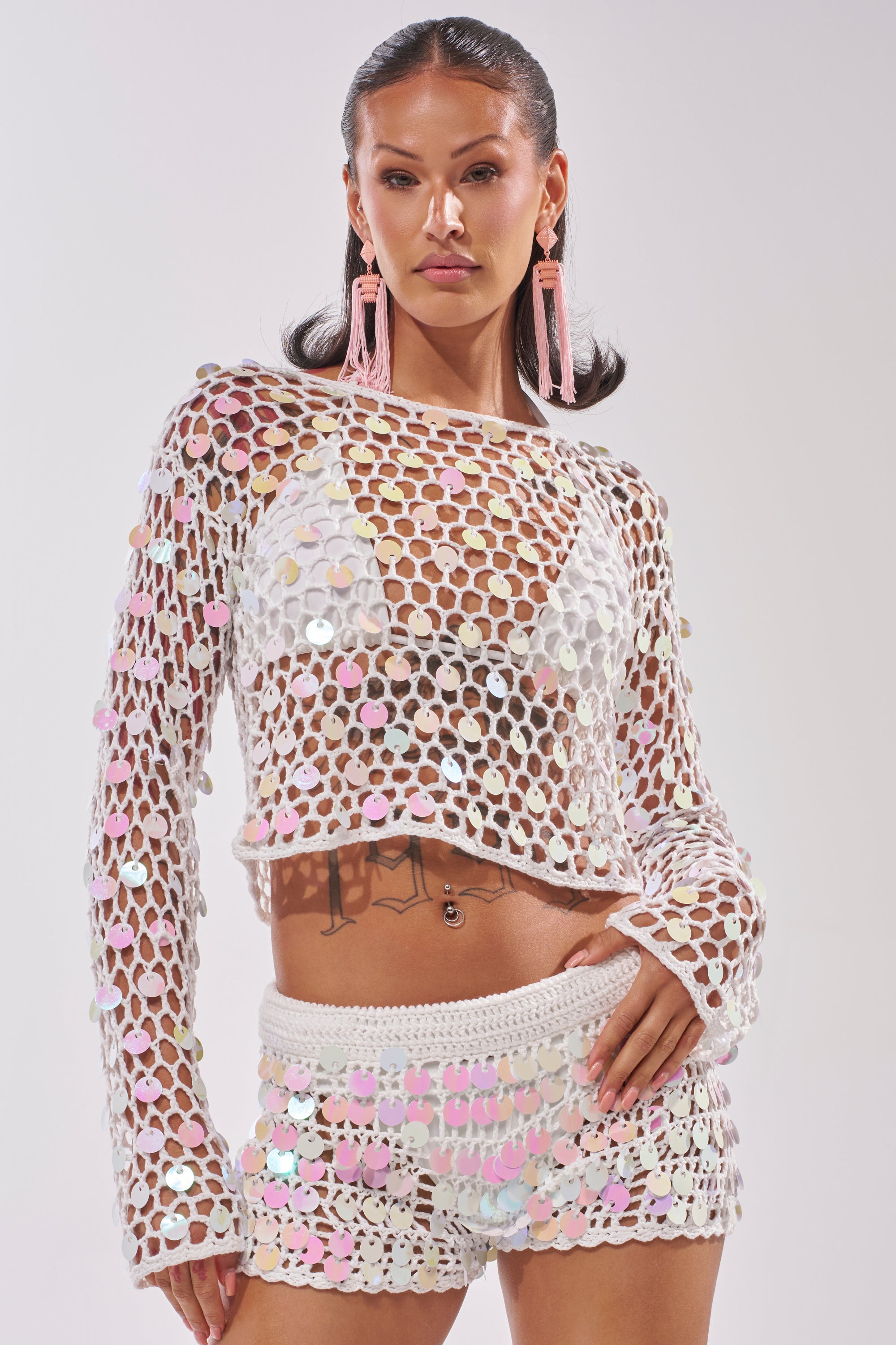 A woman poses confidently in the BEACH GIRLY CROCHET SWEATER, a sheer white crochet top adorned with large iridescent sequins, paired with matching shorts. She completes her look with pink tassel earrings and sleek, styled-back dark hair. 1