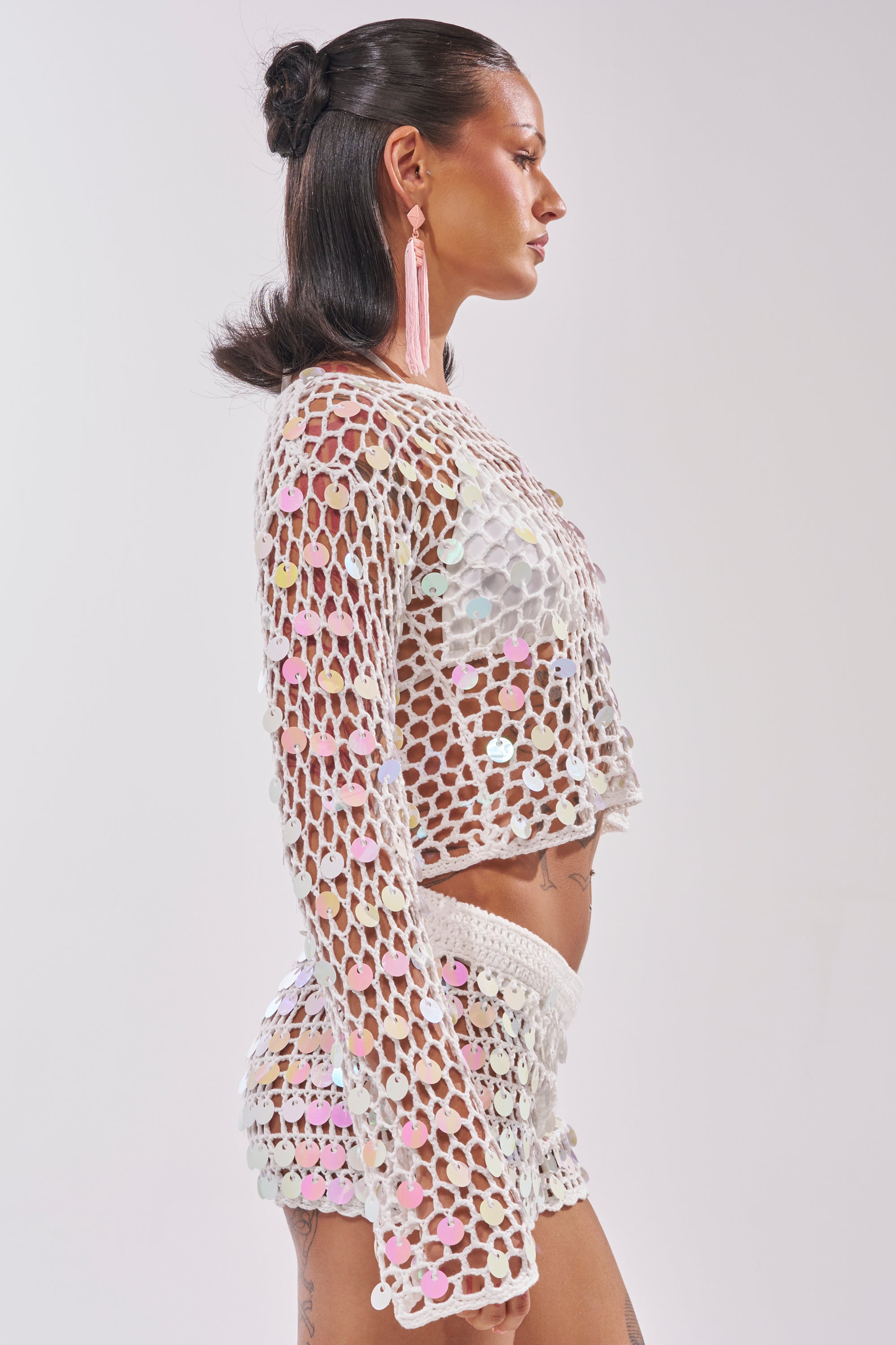 A woman with dark, slicked-back hair in a bun wears the BEACH GIRLY CROCHET SWEATER, a white sequin crochet piece paired with matching shorts. The shimmering set is see-through as she stands in profile against a white background. 4
