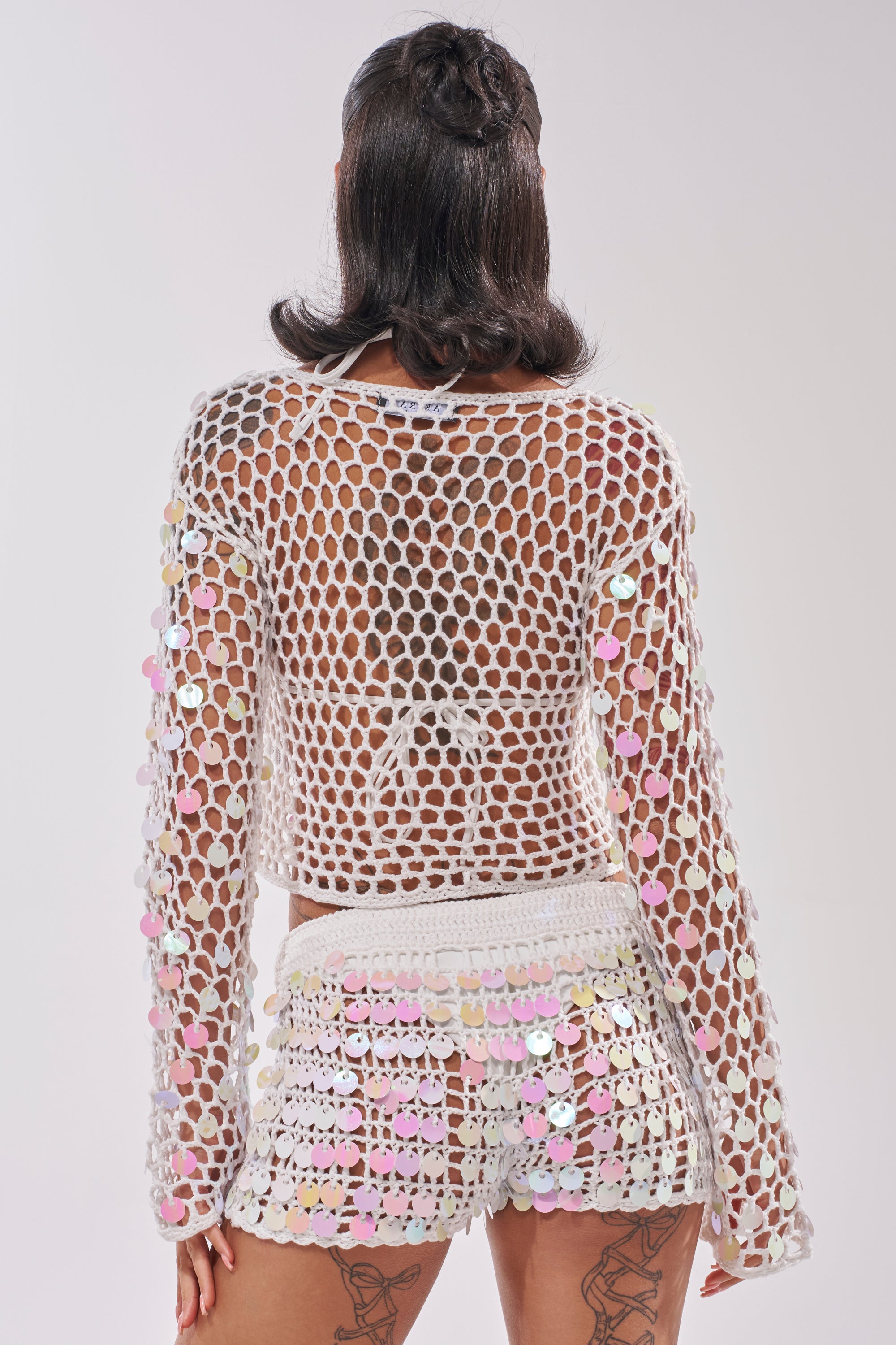 A person with shoulder-length dark hair is seen from the back, wearing the BEACH GIRLY CROCHET SWEATER in white with iridescent sequins, paired with a matching mini skirt. Tattoos are visible on their upper thighs. 5