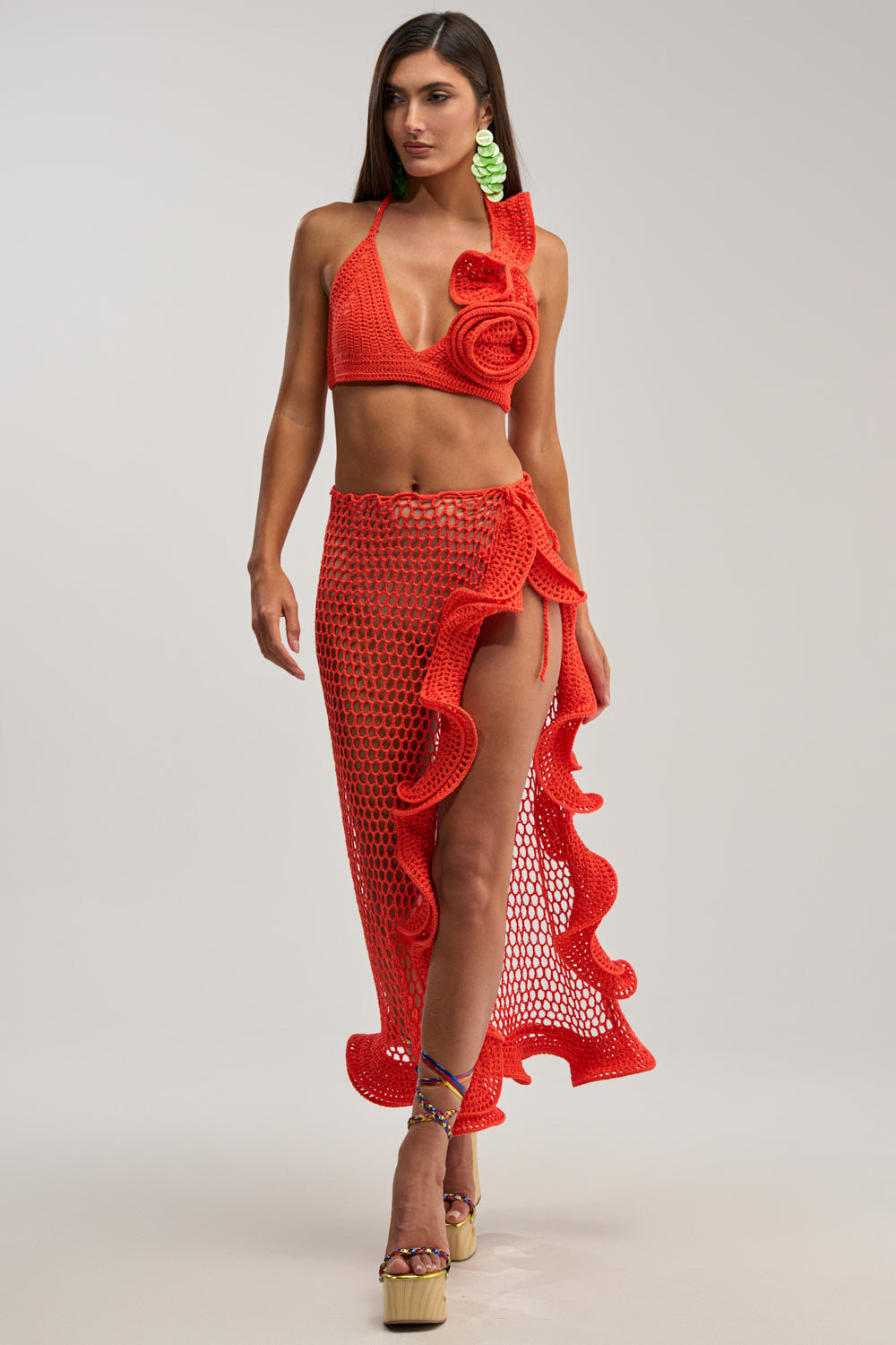A woman confidently poses in the BEACH BABE CROCHET MAXI SKIRT, featuring ruffled edges and a thigh-high slit, paired with a matching orange crochet halter top, platform sandals, and statement green earrings against a plain background.