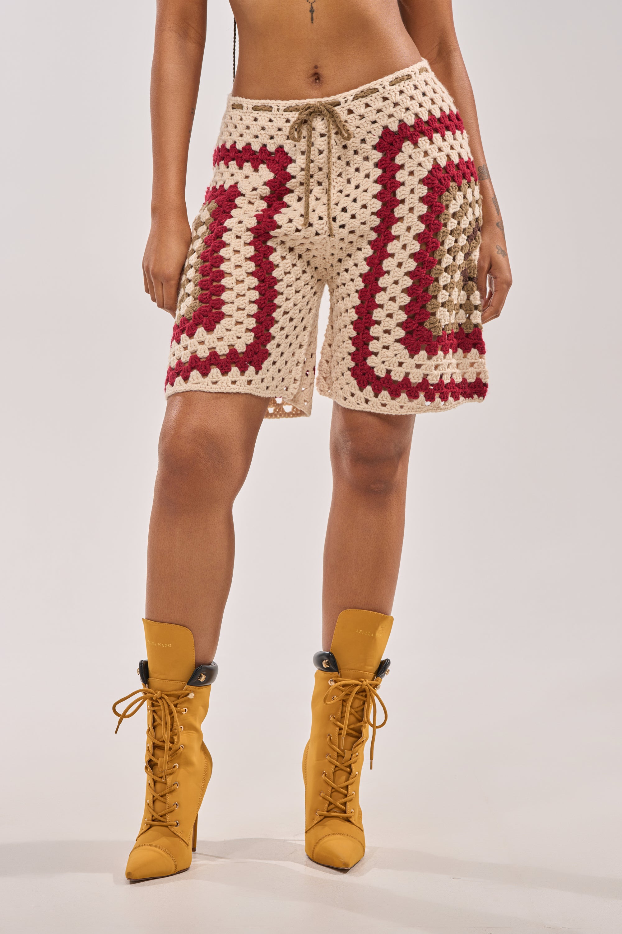 A person wearing the VACATION VIBES CROCHET SHORT—beige bermuda shorts with red, brown, and cream squares—styled with mustard lace-up heeled boots for an effortless beach resort look against a plain background. 4