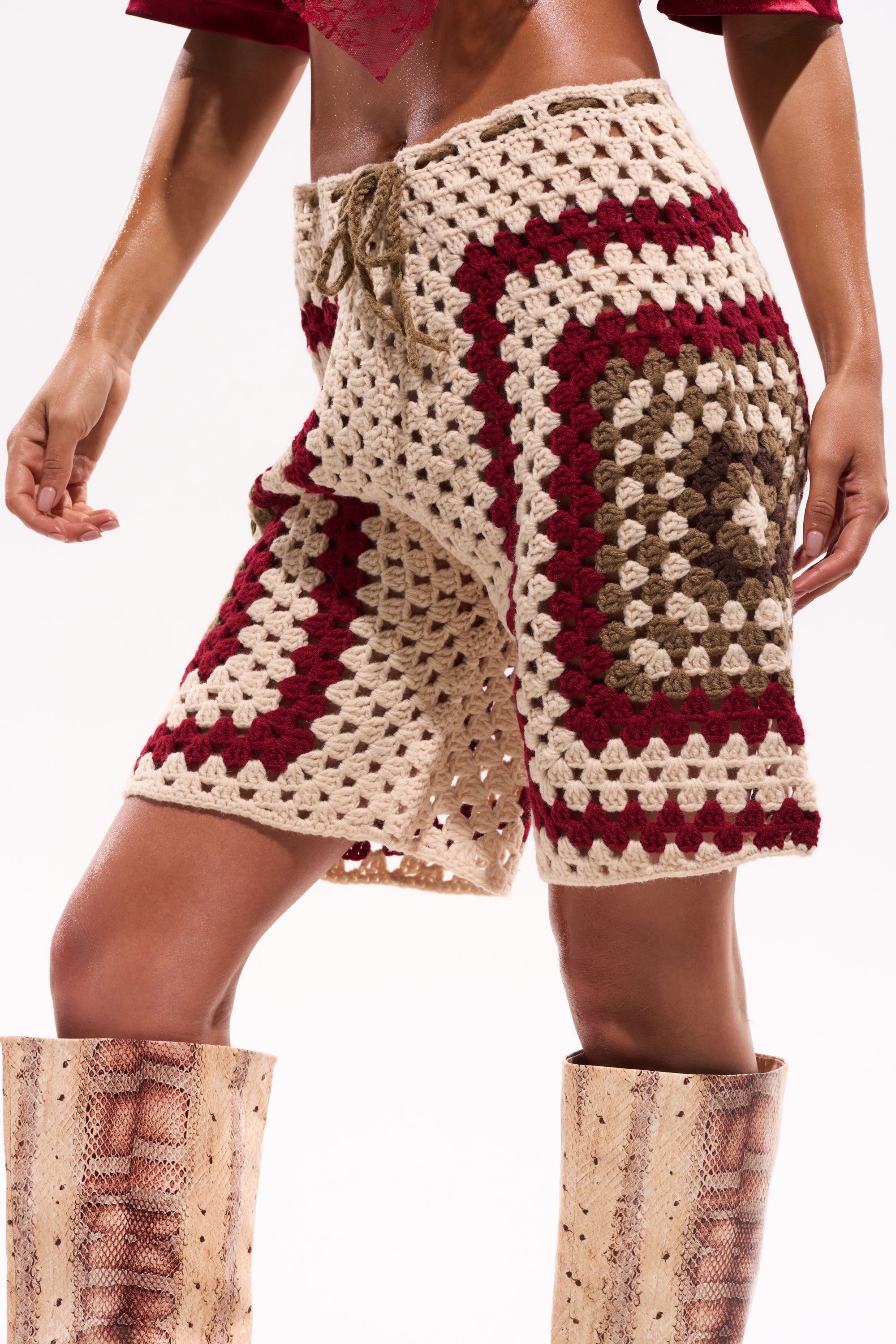 A person stands against a plain background wearing the VACATION VIBES CROCHET SHORT in beige, brown, and red with a drawstring, styled with tall tan snakeskin boots and a cropped red top. 0