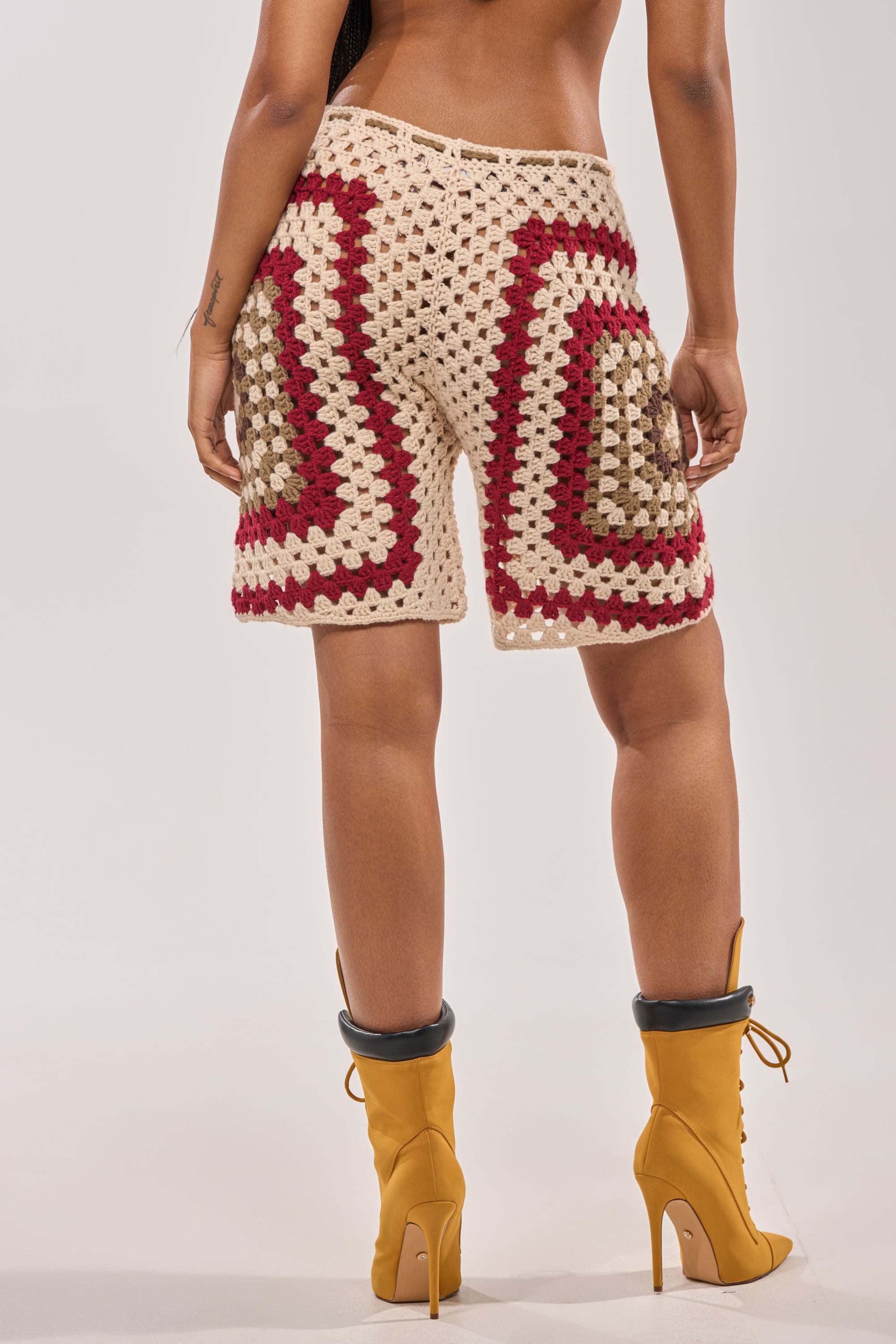 A person faces away, showcasing their back in VACATION VIBES CROCHET SHORTS—beige, red, and brown—styled with mustard-yellow heeled boots against a plain light background for an eye-catching beach resort look. 7