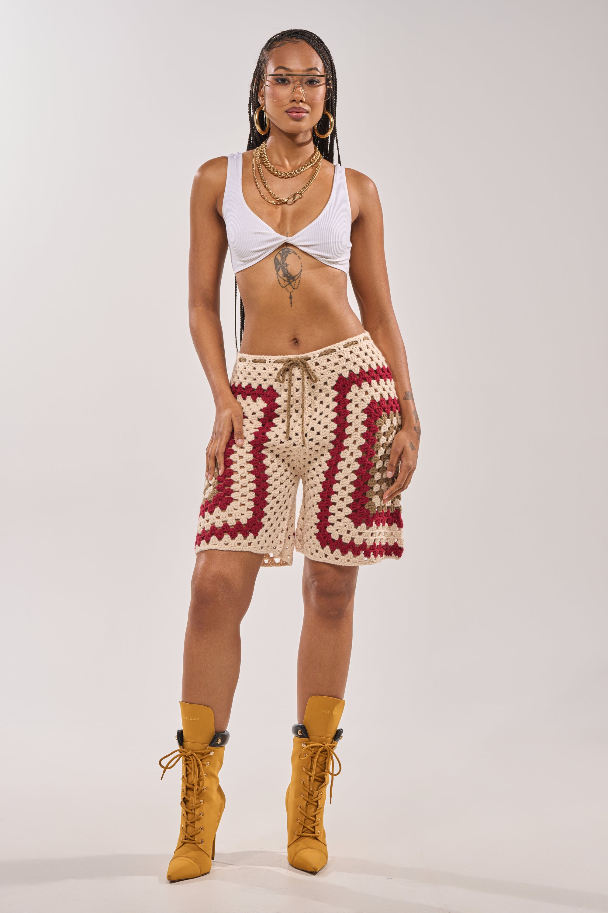 A woman with braided hair wears the VACATION VIBES CROCHET SHORT in beige and red, a white crop top, yellow lace-up boots, gold jewelry, and glasses—creating an effortless beach resort look against a plain background. 8
