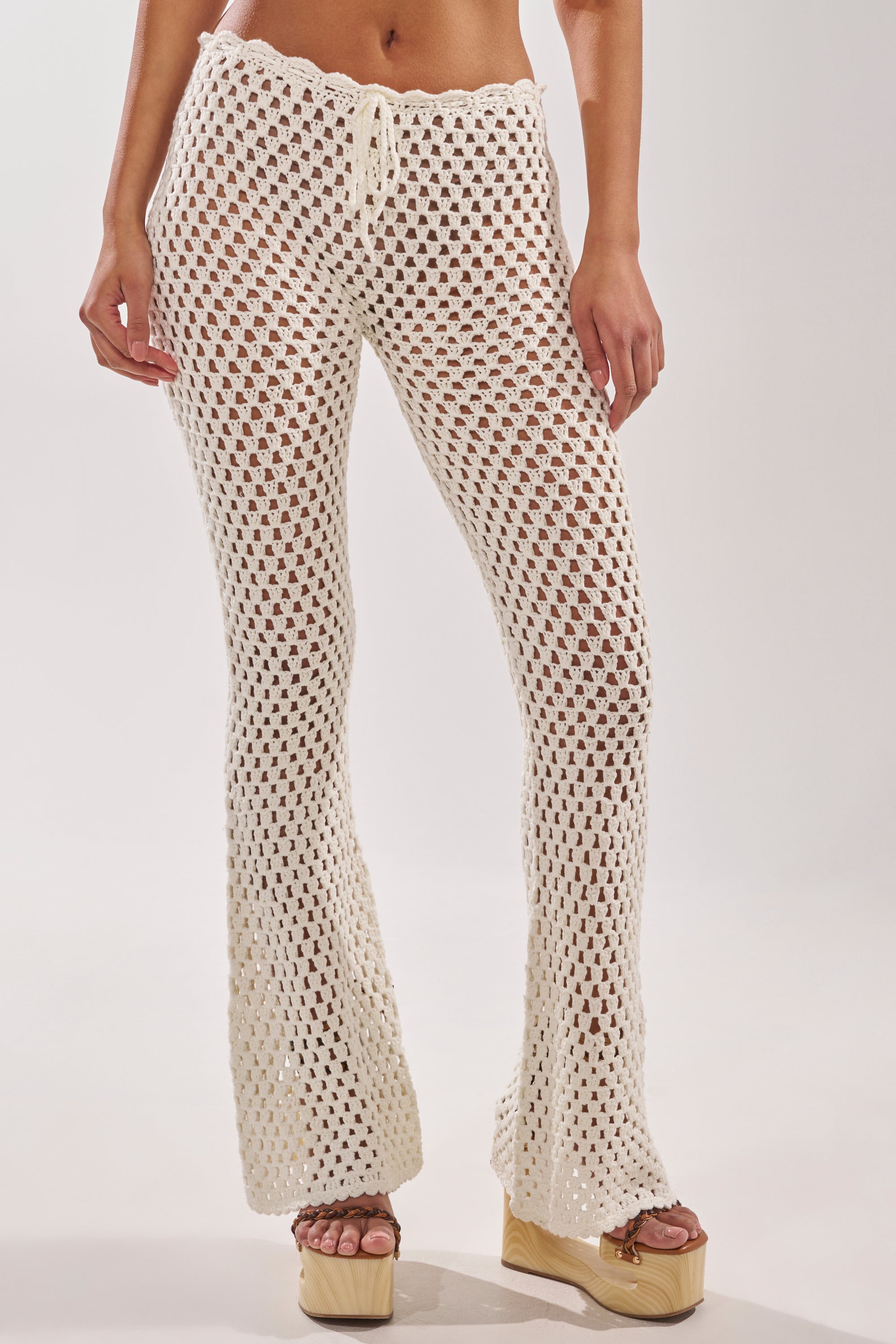 A person models the BEACH VIBES CROCHET FLARED PANT—high-waisted, white, semi-sheer with a drawstring waist and flared legs—styled for resort wear with platform sandals against a light background. 1