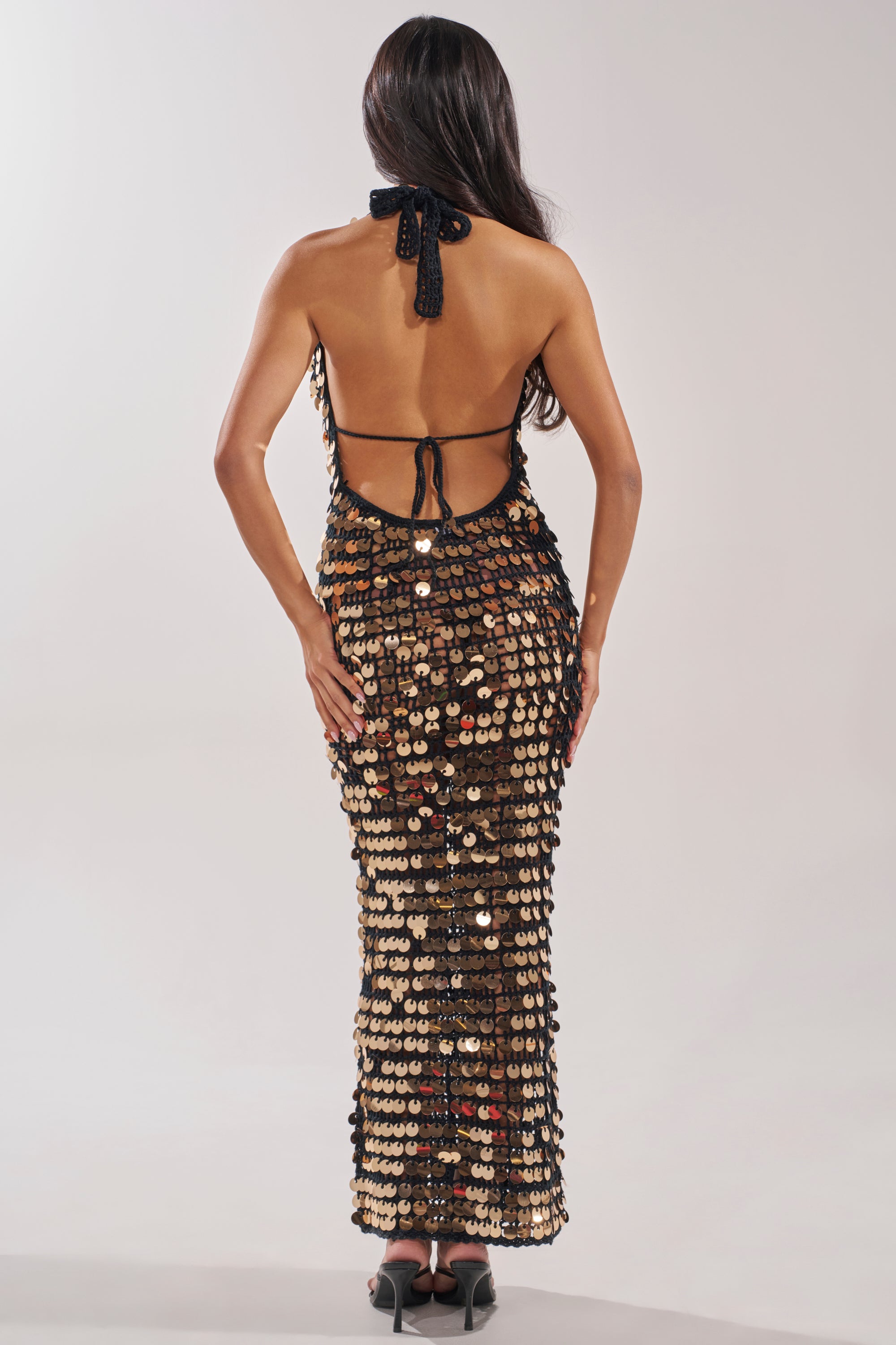 A woman with long dark hair faces away, wearing the ARIEL SEQUIN CROCHET MAXI DRESS IN BLACK, covered in shiny gold and silver sequins, paired with black high-heeled sandals against a plain light background. 4