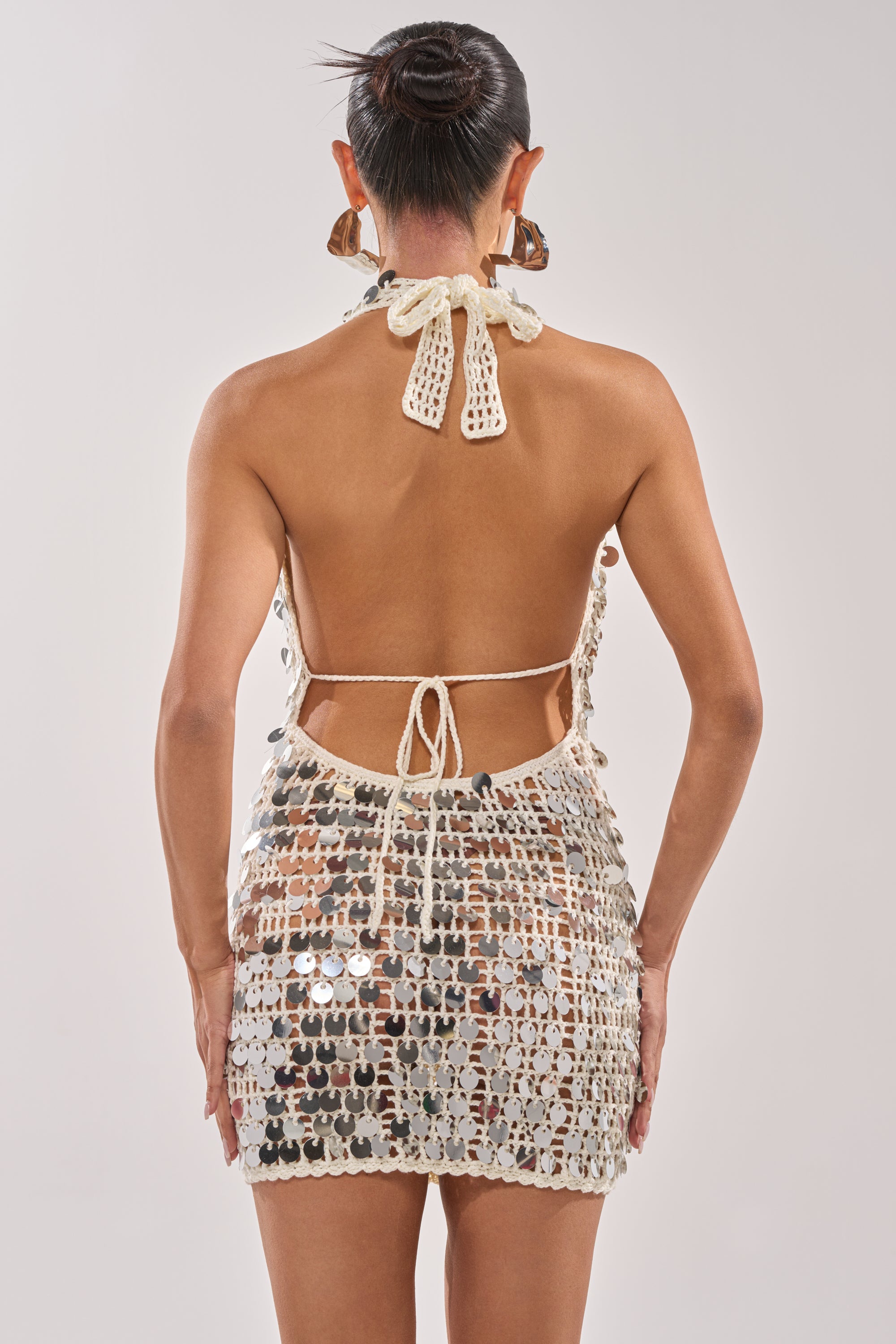 A person faces away, wearing the ARIEL SEQUIN CROCHET MINI DRESS IN WHITE—a disco-inspired halter mini with tie-back and neck. Their hair is in a sleek bun, finished with large statement earrings. 4