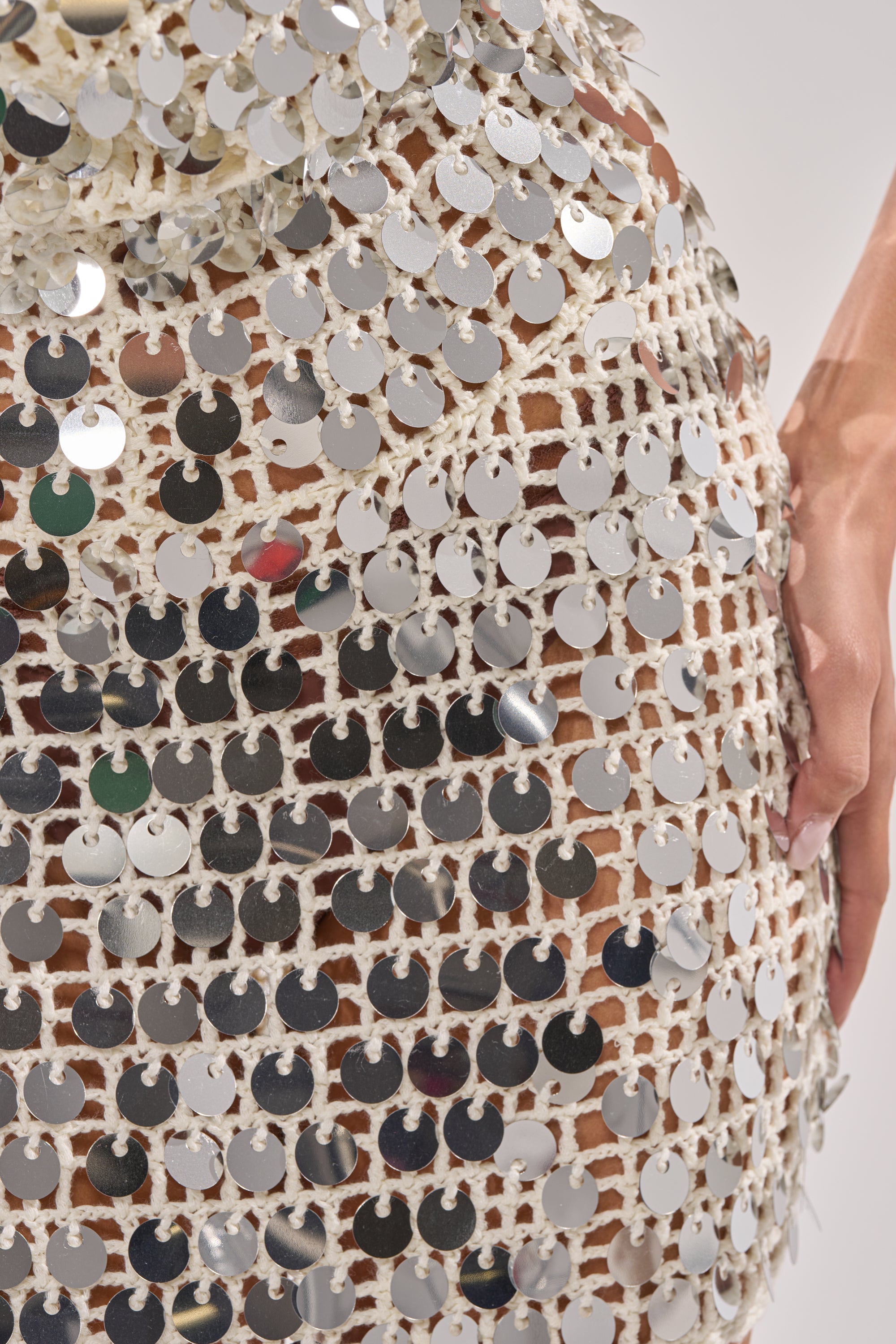 Close-up of a person wearing the ARIEL SEQUIN CROCHET MINI DRESS IN WHITE, featuring cream crochet with large silver sequins that catch the light for a dazzling, disco-inspired look. Their hand rests by their side. 6