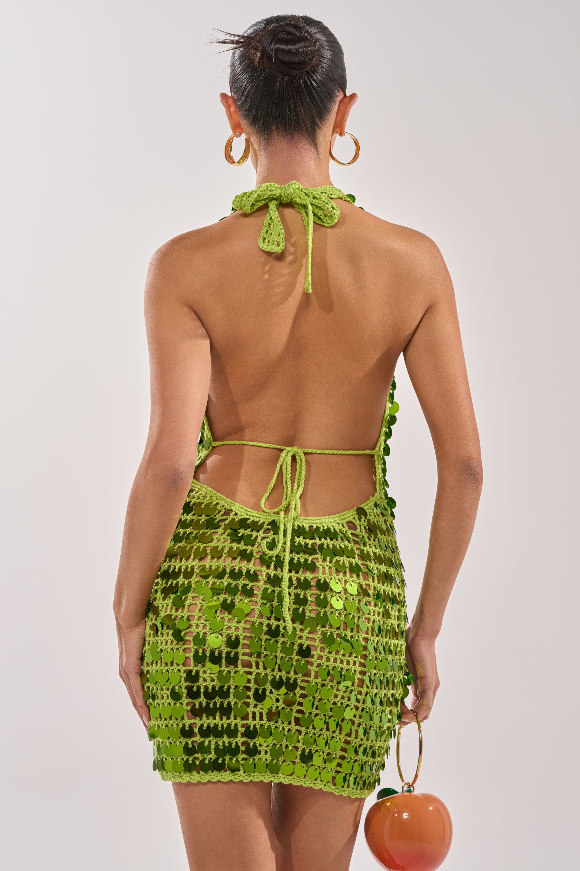 A woman with her hair in a bun wears the ARIEL SEQUIN CROCHET MINI DRESS IN GREEN, featuring a backless halter style with thin ties. She holds a round orange purse, wears large gold hoop earrings, and stands against a plain light background. 6