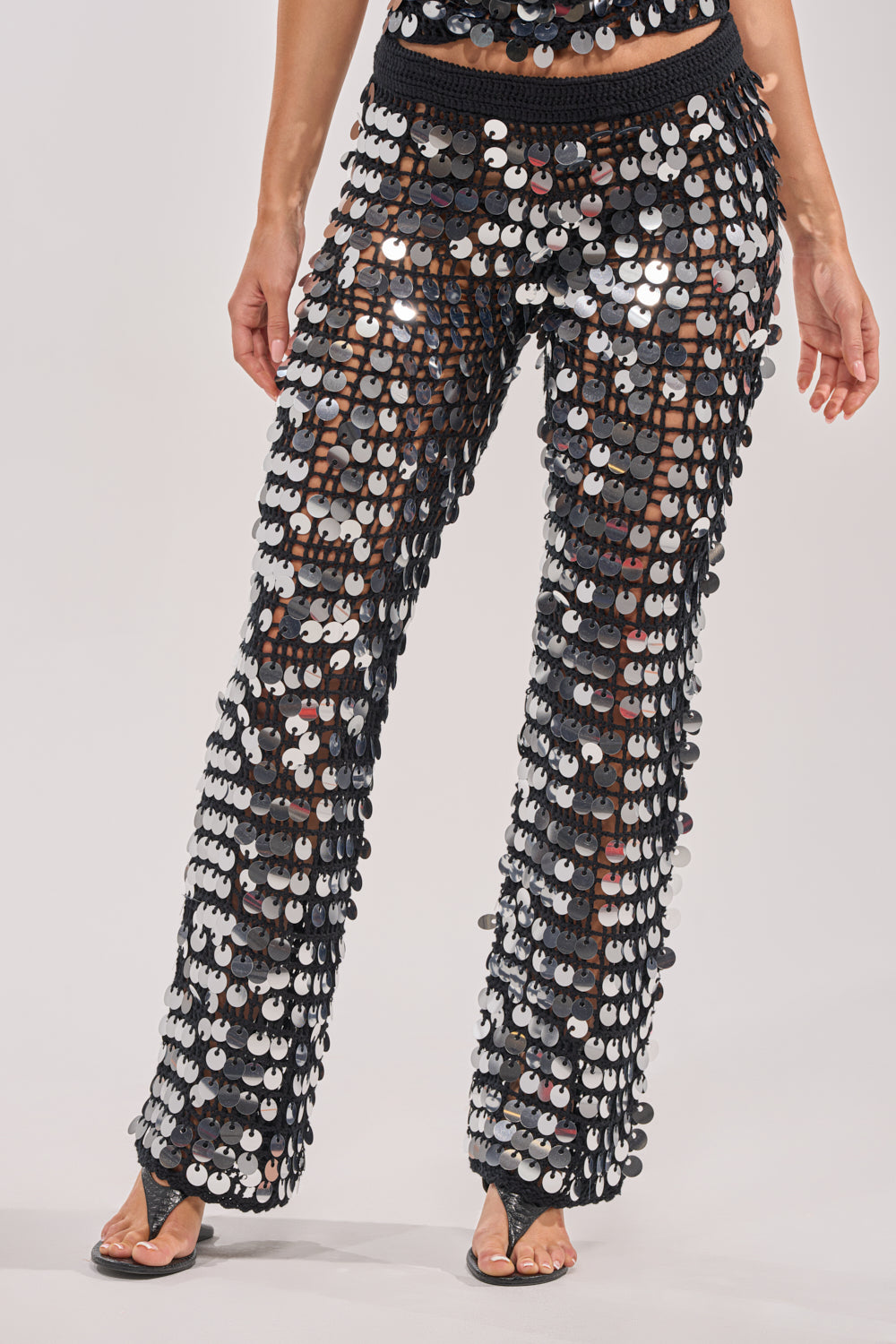 A person models the GIVE IT TO ME BABY CROCHET PANT—black flare festival pants decorated with large reflective silver and black sequins—paired with black sandals against a plain background. 1