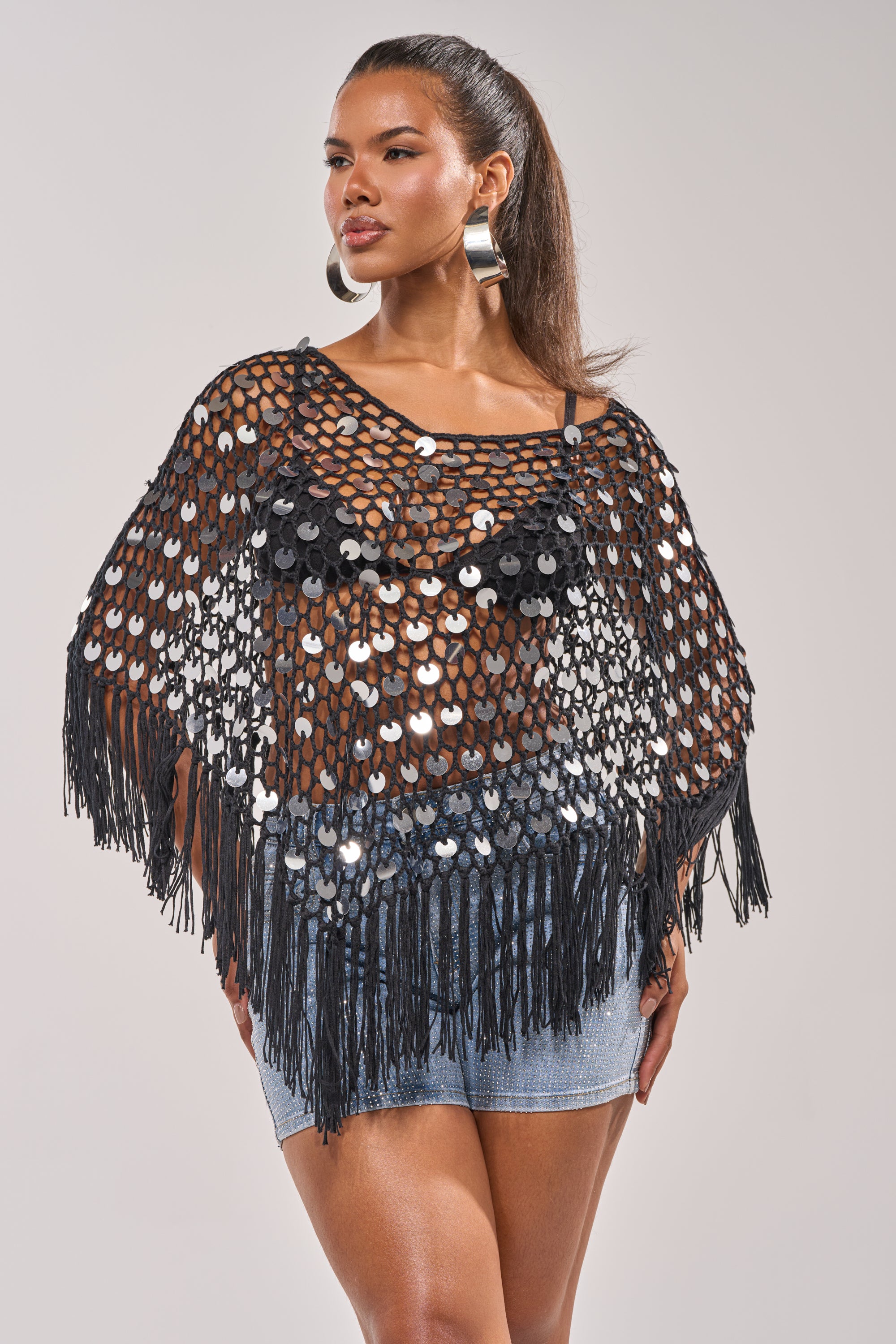 She wears the UNDER THE LIGHTS CROCHET SEQUIN PONCHO over a black top and denim skirt for a standout festival look, finishing her summer style with hoop earrings and a high ponytail. 0