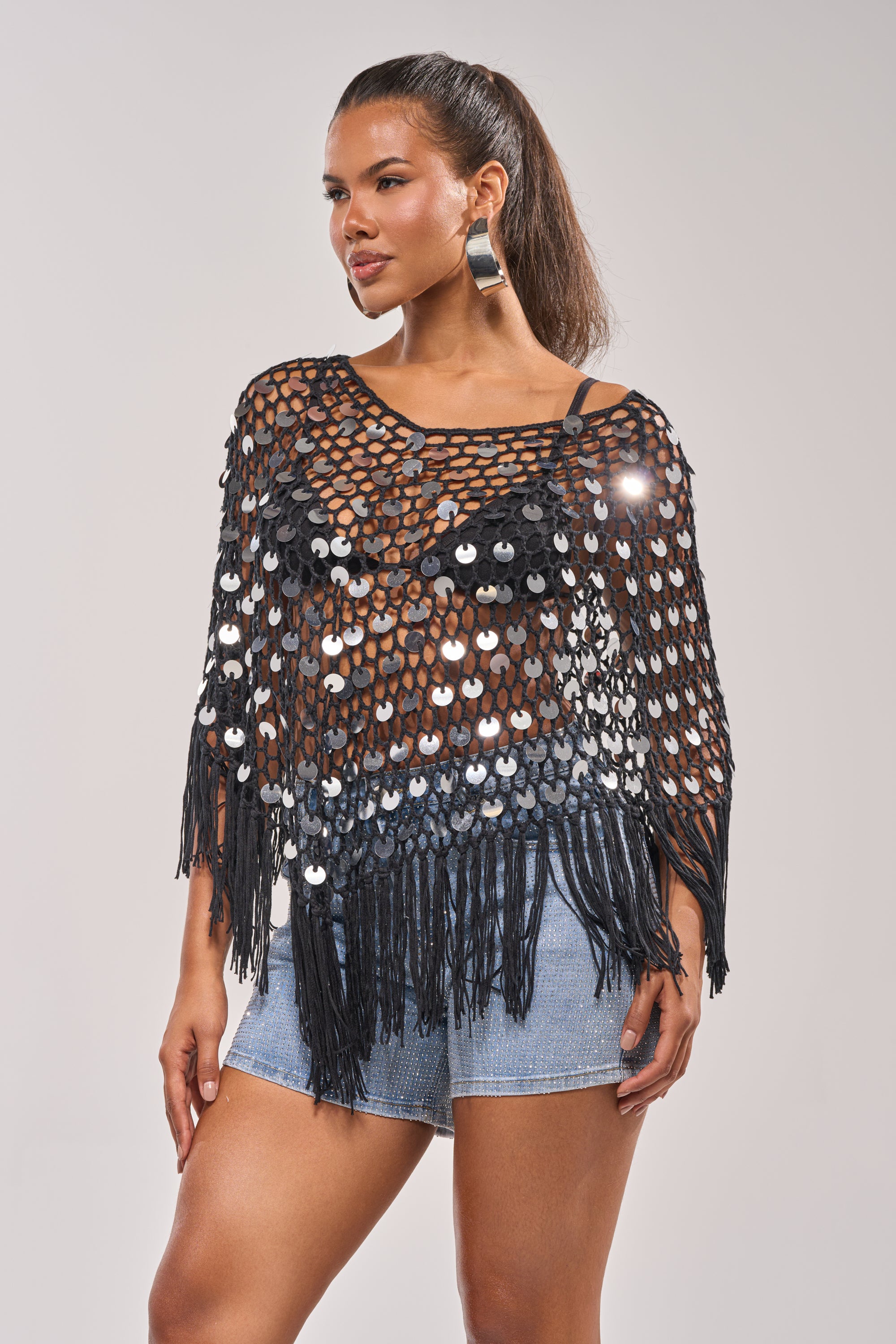 Wearing the UNDER THE LIGHTS CROCHET SEQUIN PONCHO over a tank and denim shorts, a woman with a ponytail and hoop earrings poses confidently against a plain background—a perfect summer festival look. 1