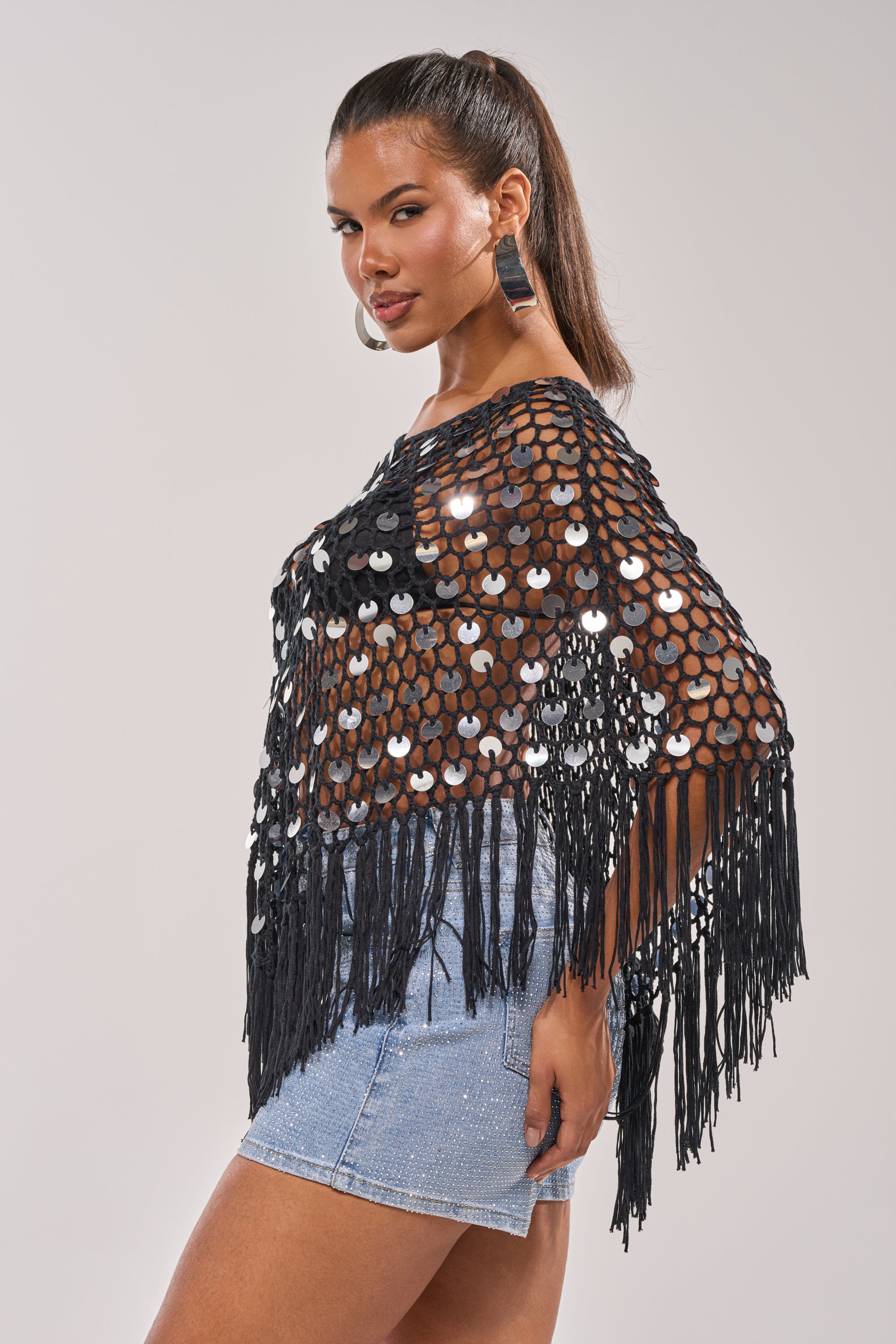 A woman models the UNDER THE LIGHTS CROCHET SEQUIN PONCHO with light blue denim shorts, her long hair in a high ponytail and large hoop earrings—set against a simple background for festival outfit inspiration. 2