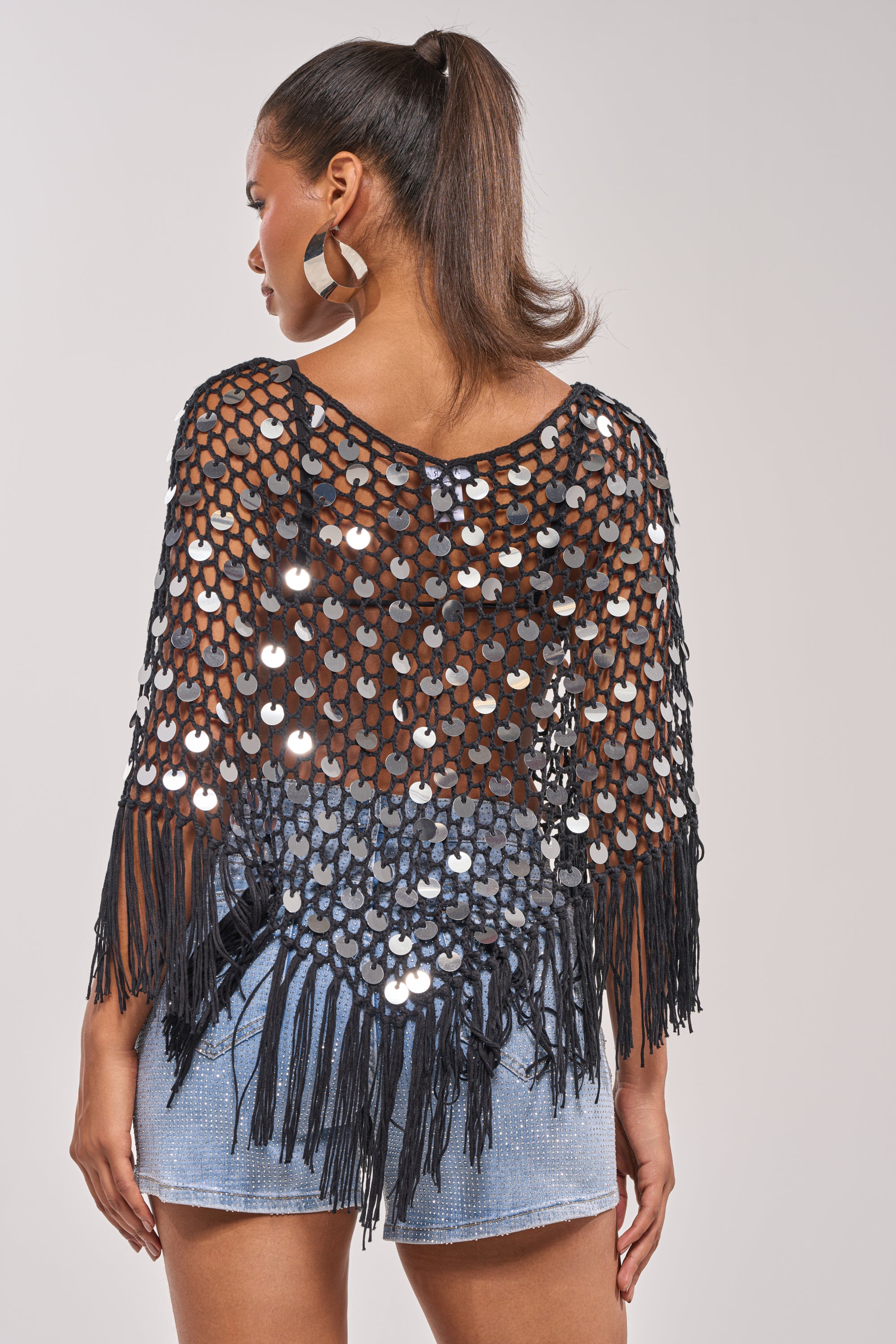 A woman with a high ponytail and large hoop earrings wears the UNDER THE LIGHTS CROCHET SEQUIN PONCHO and a light blue mini skirt, facing away from the camera against a plain background. Perfect for festival outfits. 3